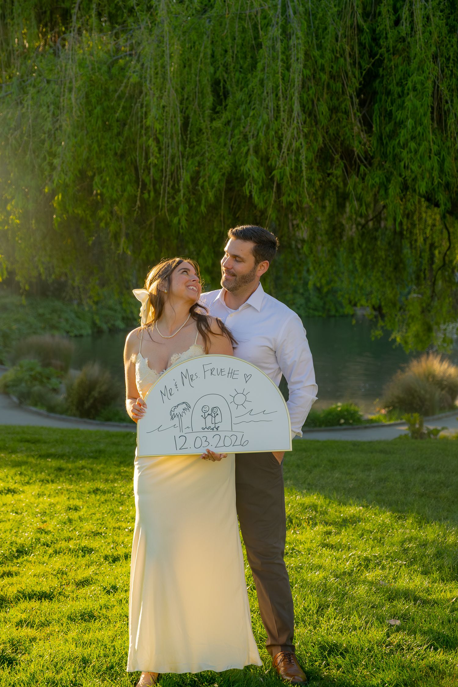 Engaged couple holding a save the date sign under a willow tree in golden hour sunlight.