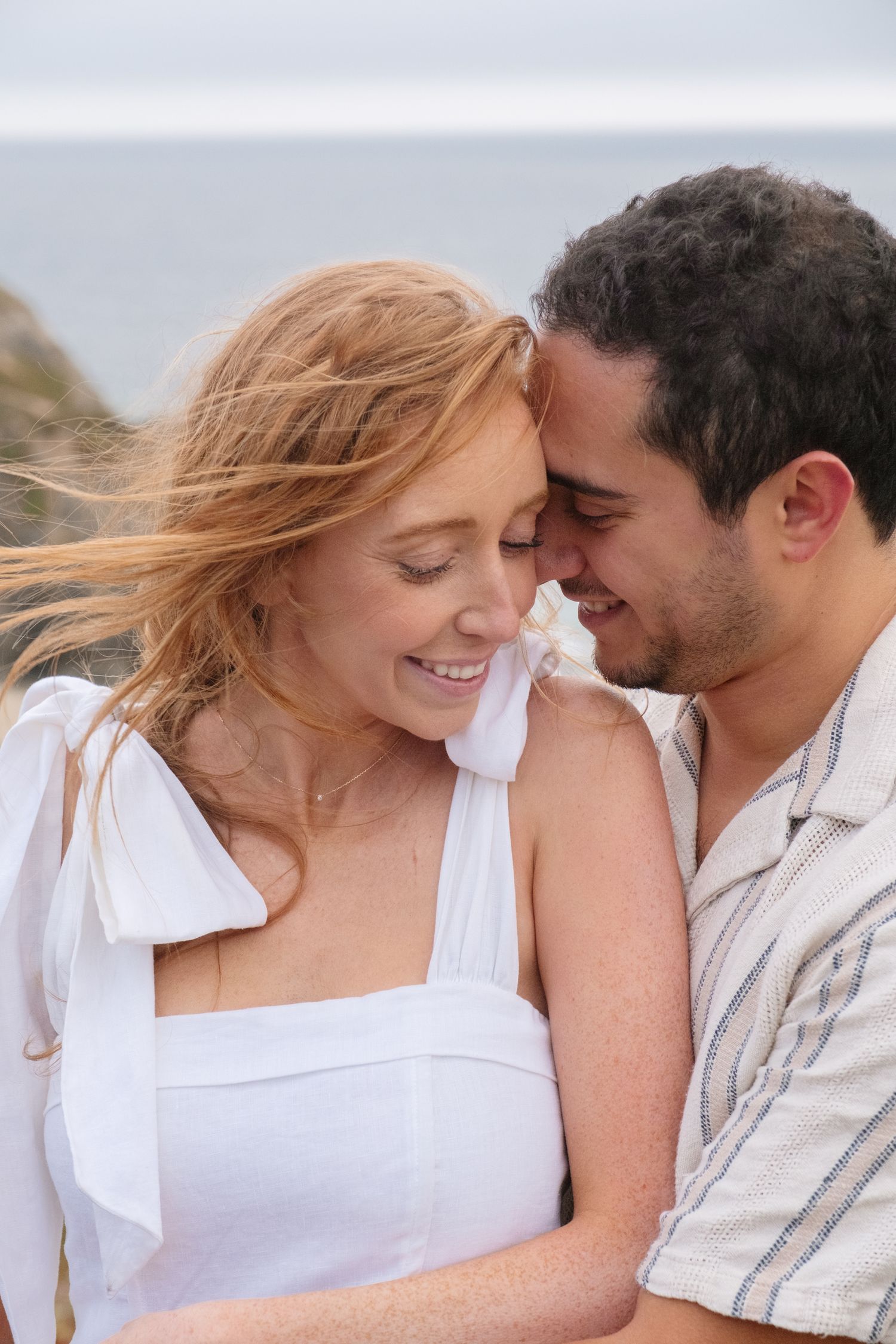 A smiling couple shares a romantic moment by the ocean, the woman in a white dress with windswept red hair.