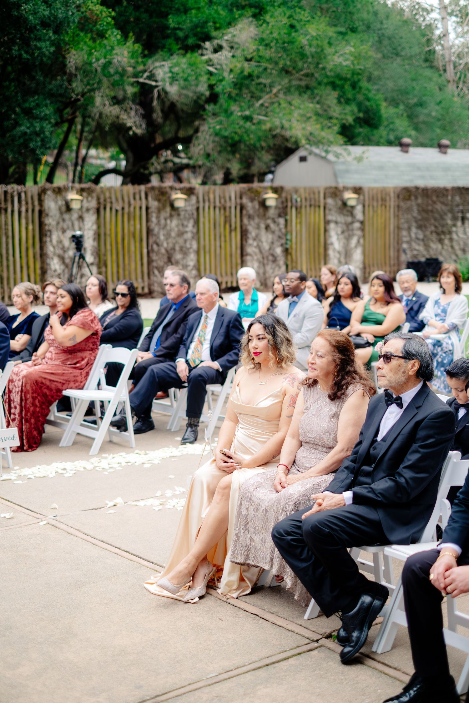 Guests in elegant attire watch outdoor wedding ceremony from white folding chairs.