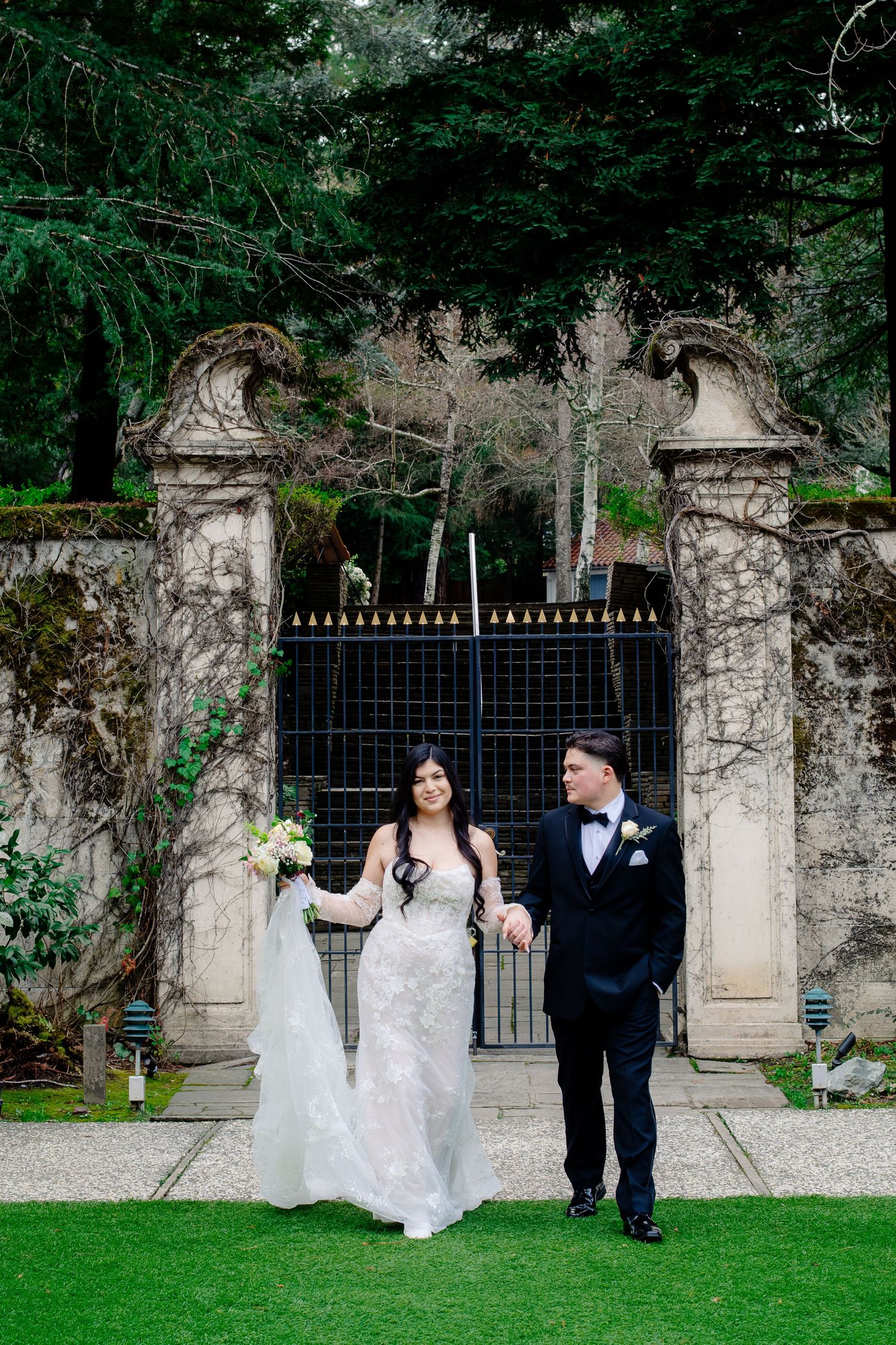 Bride and groom walk arm in arm before stone gate surrounded by lush greenery.