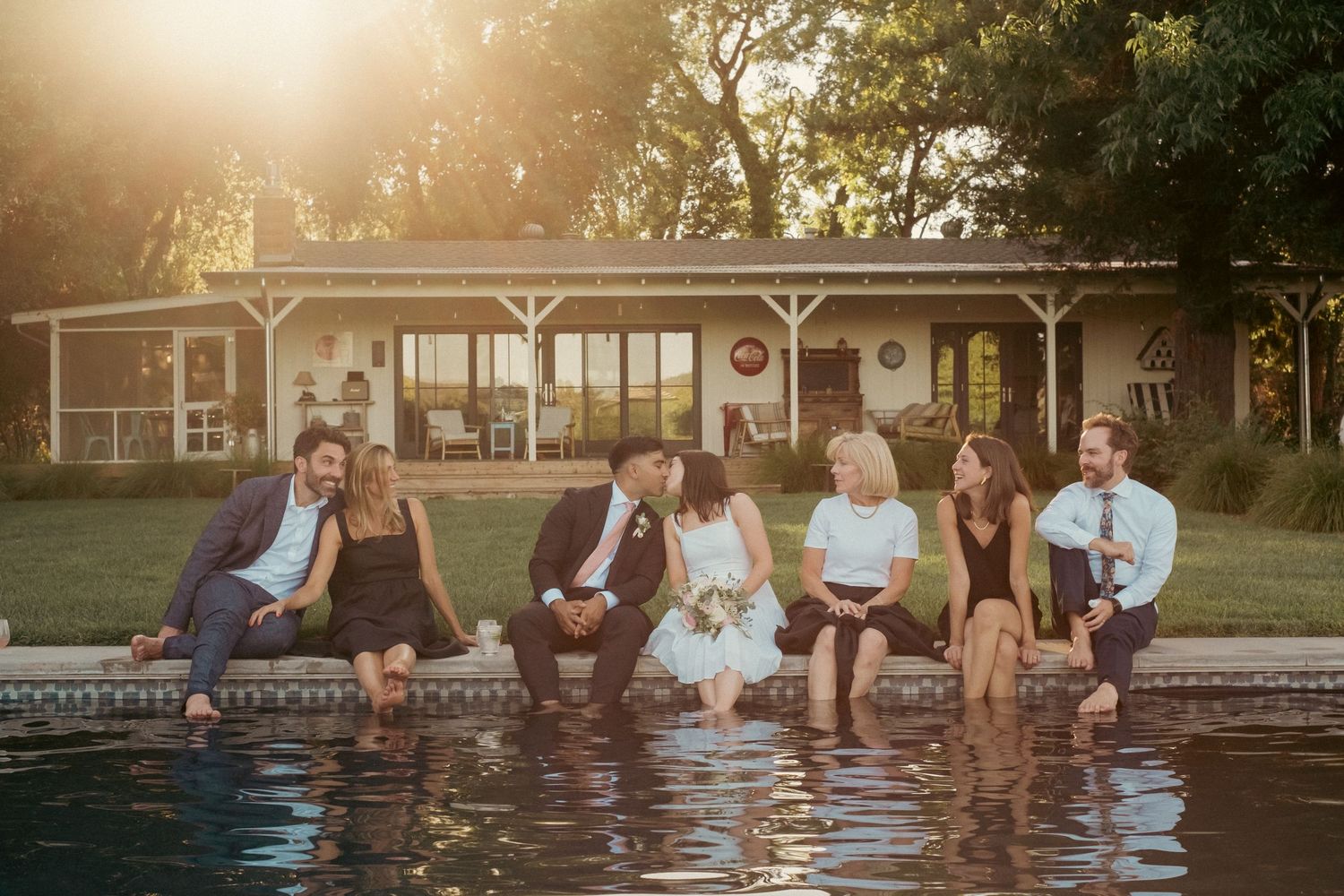 Wedding party sits with feet in pool at golden hour beside a charming country house.