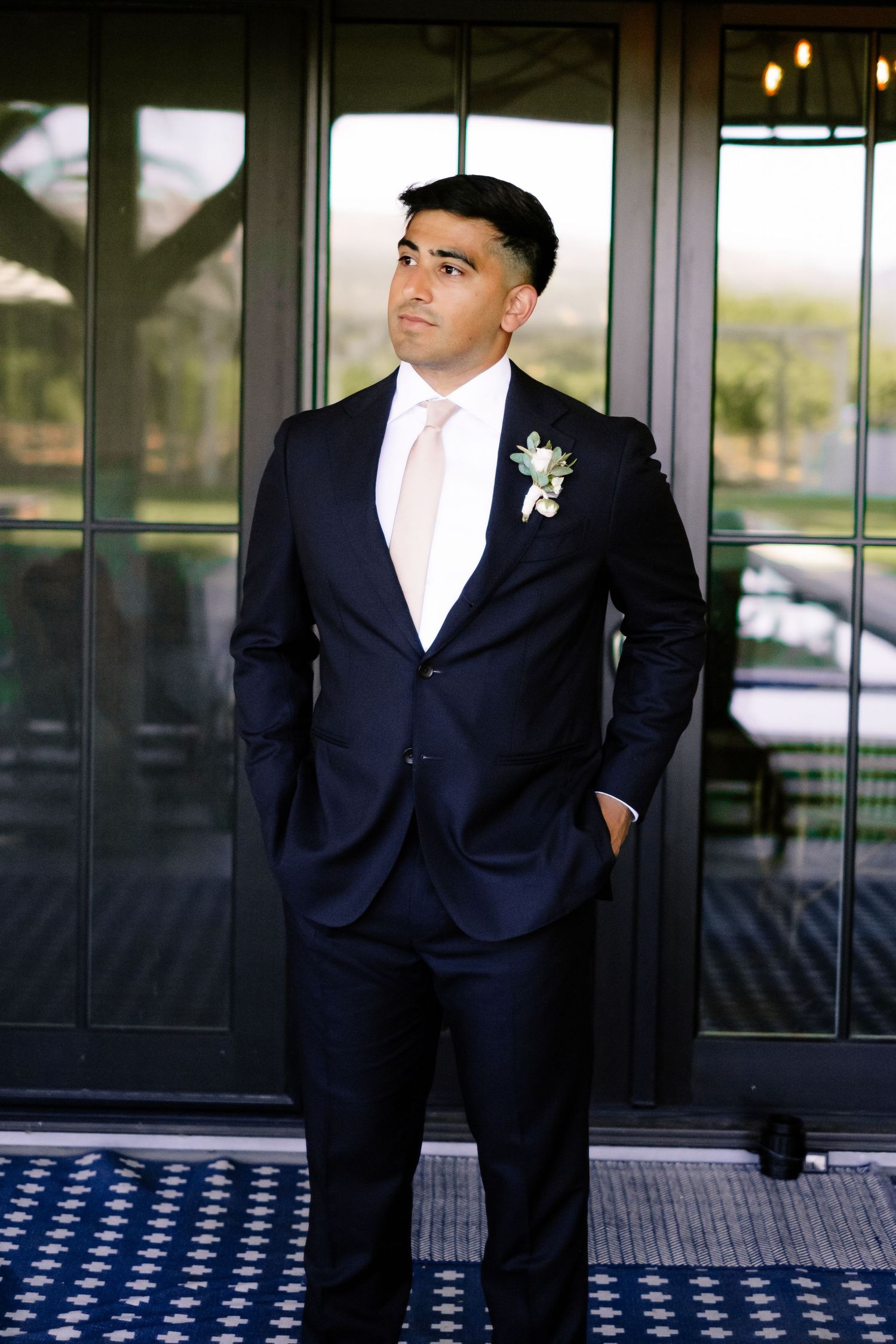 Groom in navy suit and blush tie with boutonniere posing before glass doors.