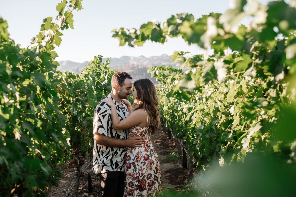 Couple embracing romantically in a lush vineyard with green grapevines and mountain backdrop on a sunny day.