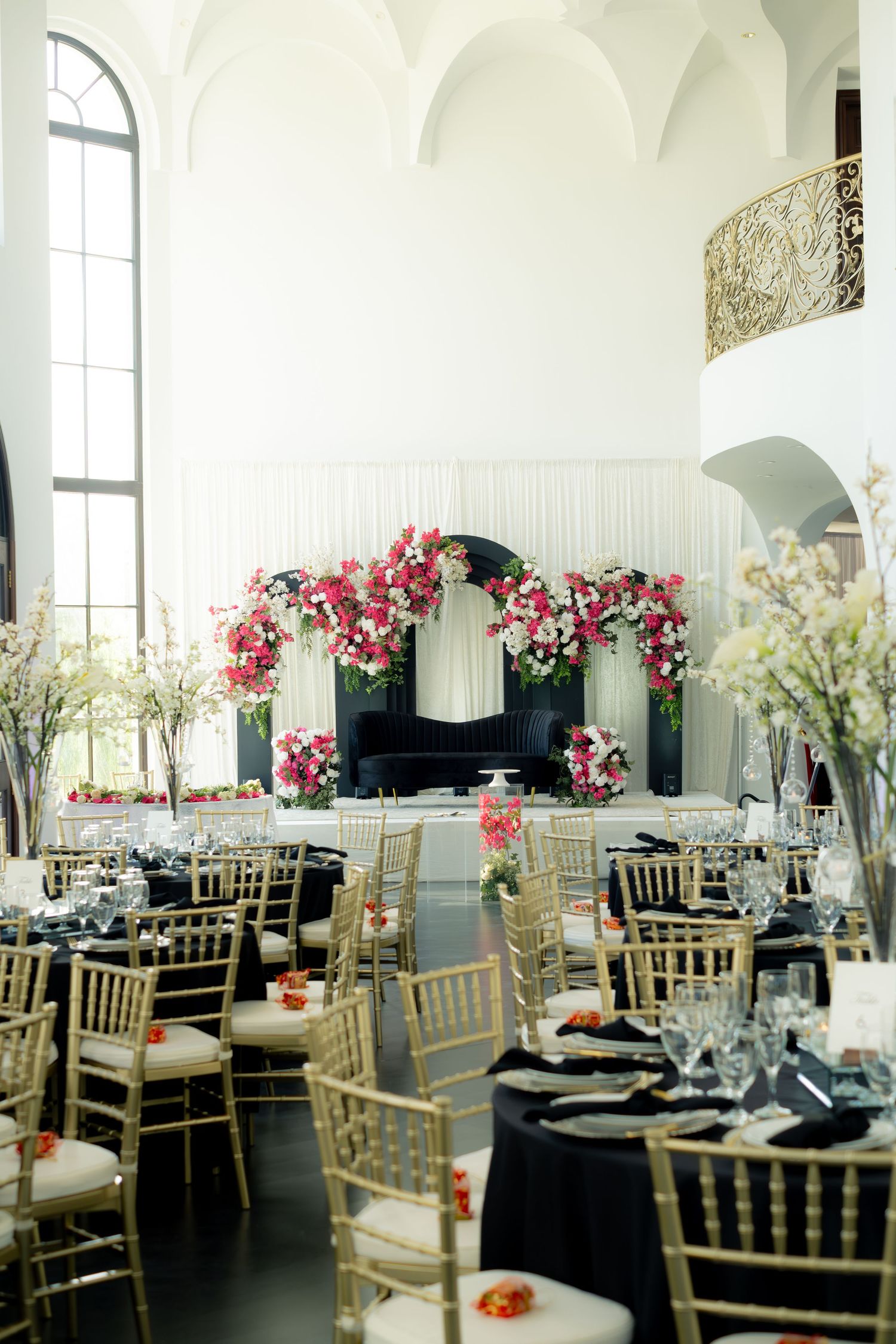 Elegant wedding reception hall with gold chiavari chairs, black linens, and pink floral arch centerpiece.