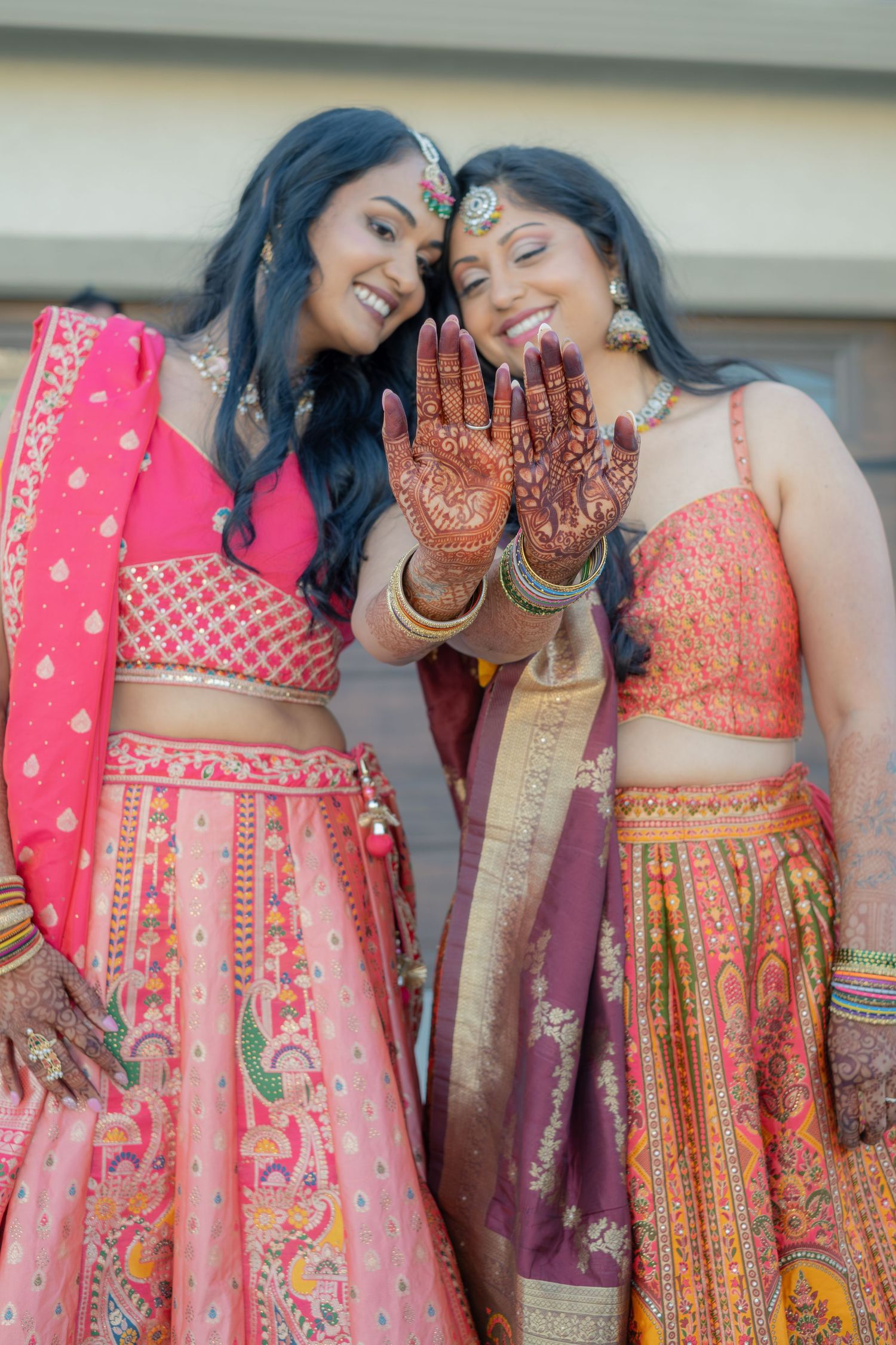Two Indian women in vibrant pink and orange lehengas smiling and showing henna hands at wedding.