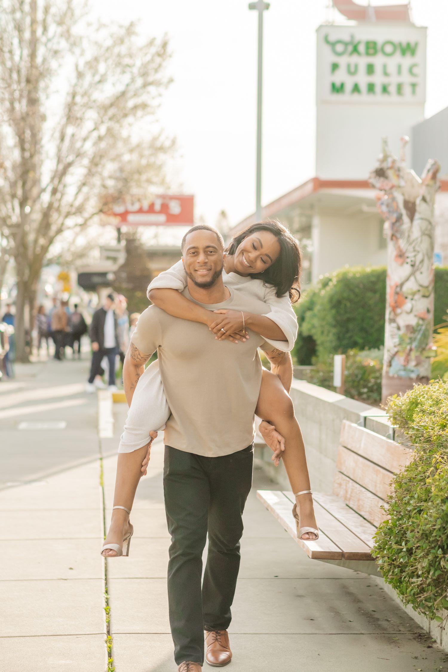 A joyful Black man gives his smiling partner a piggyback ride near Oxbow Public Market on a sunny day.