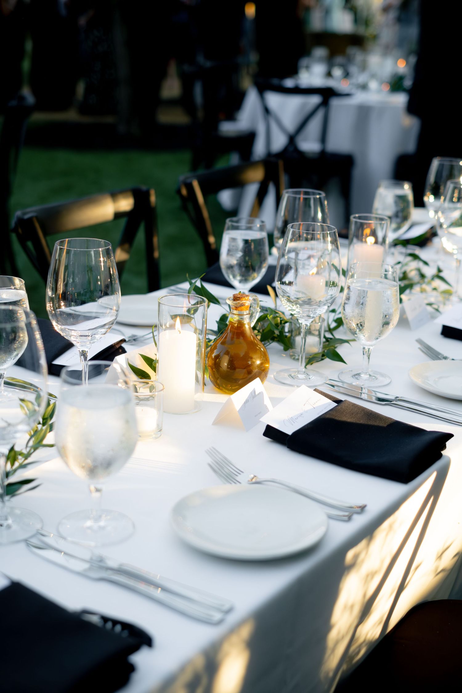 Elegant wedding reception table set with white linens, black napkins, crystal glassware, candles, and olive branch greenery.