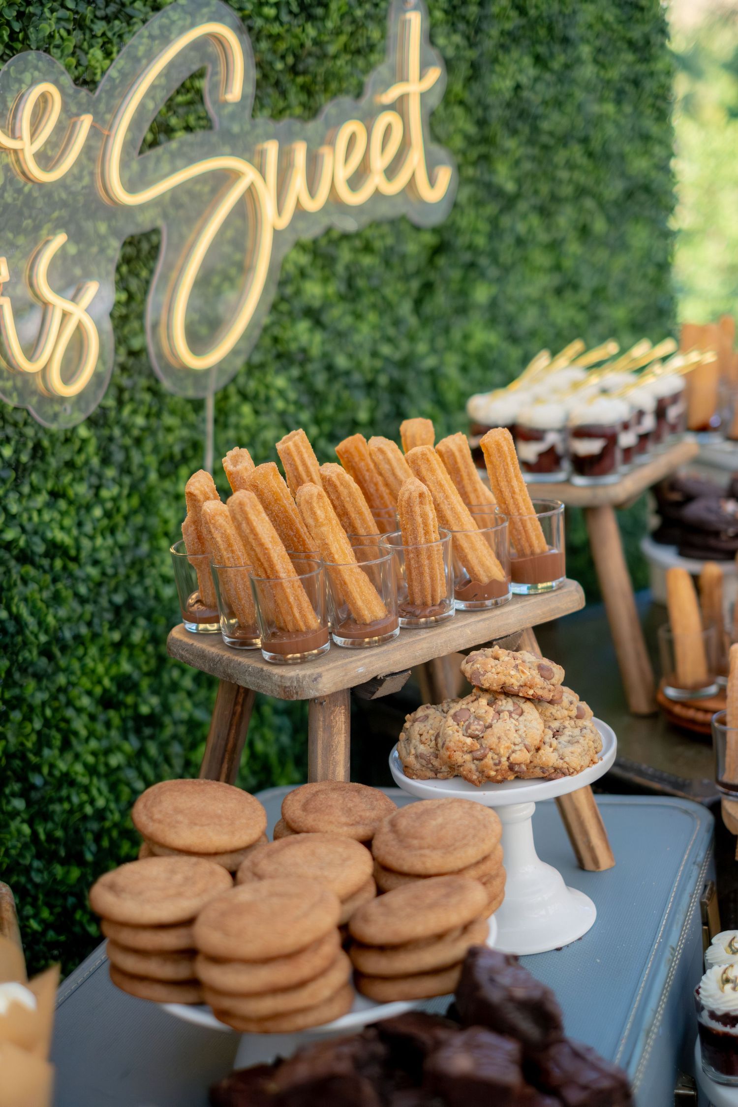Wedding dessert table featuring churros, cookies, and sweets with a neon Sweet sign backdrop.