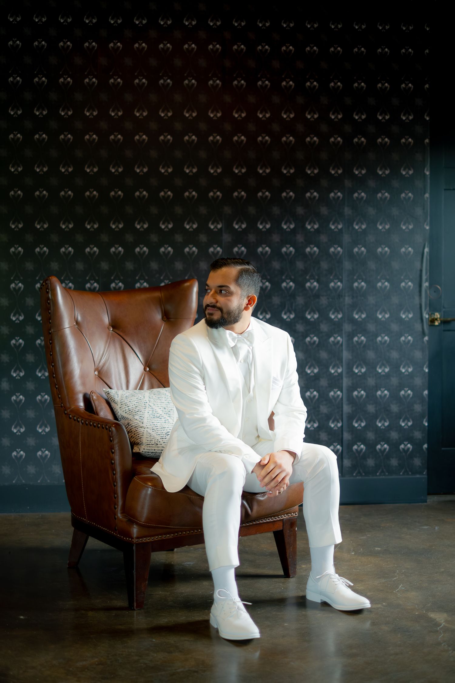 Groom in white suit sitting elegantly in a brown leather chair against a dark patterned wall.