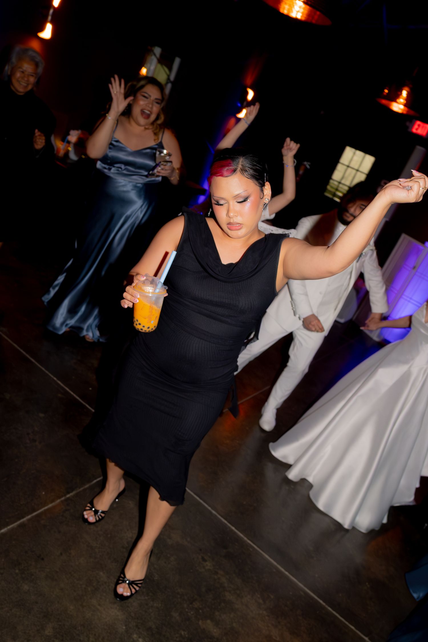 Guest in black dress dances energetically holding a drink at a lively wedding reception.