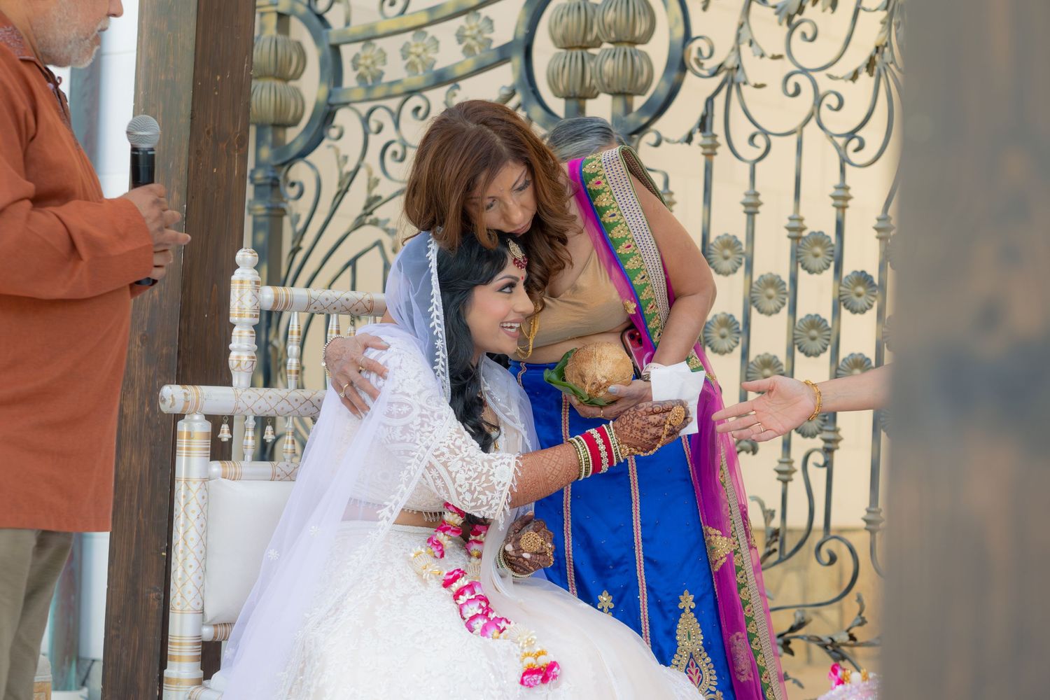 Indian bride in white lehenga receives blessings during traditional Hindu wedding ceremony at ornate gate.