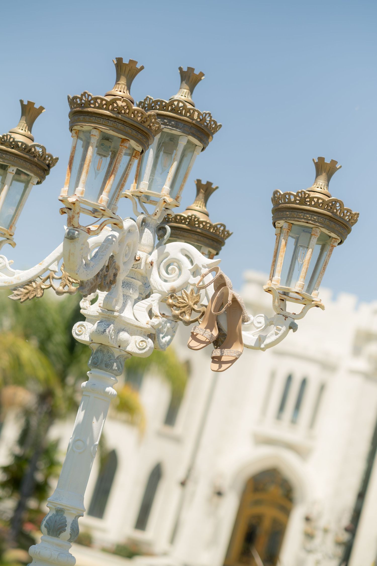 Elegant ornate white and gold street lamp post with bridal heels hanging, wedding venue in background.
