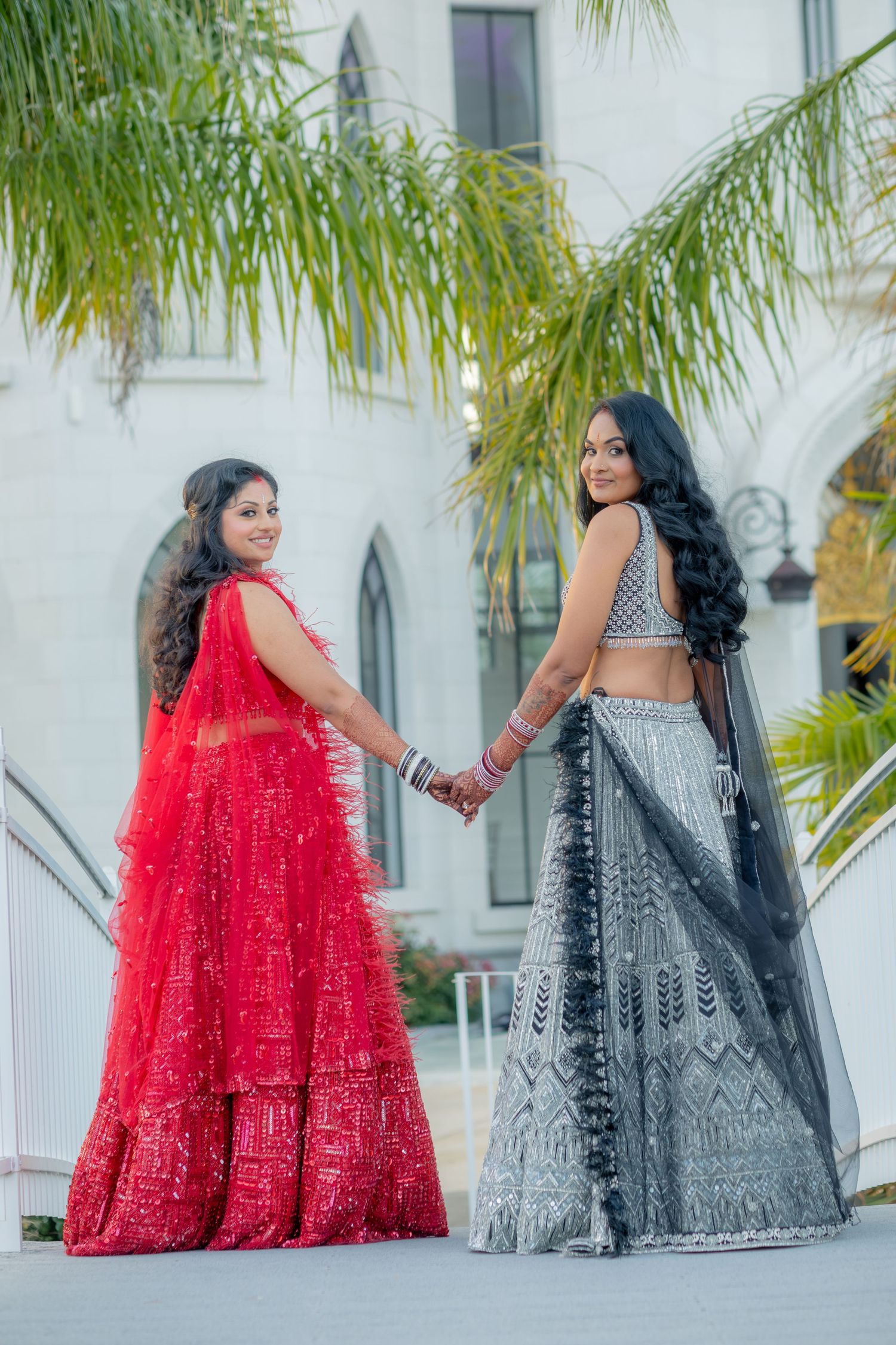 Two women in red and silver Indian lehengas hold hands while posing outside a white building.