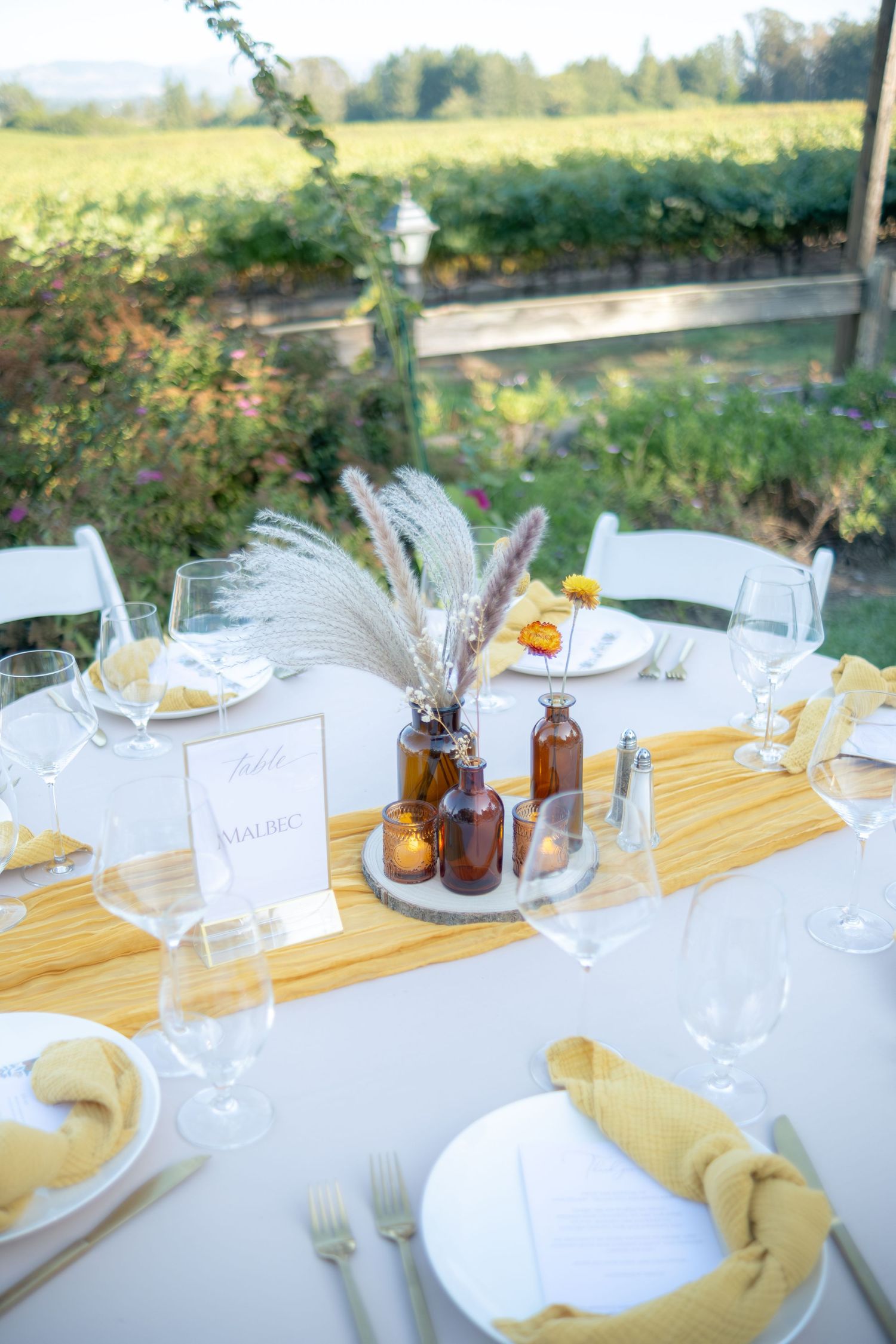 Elegant outdoor wedding reception table with yellow runner, amber bottles, pampas grass centerpiece, and vineyard backdrop.