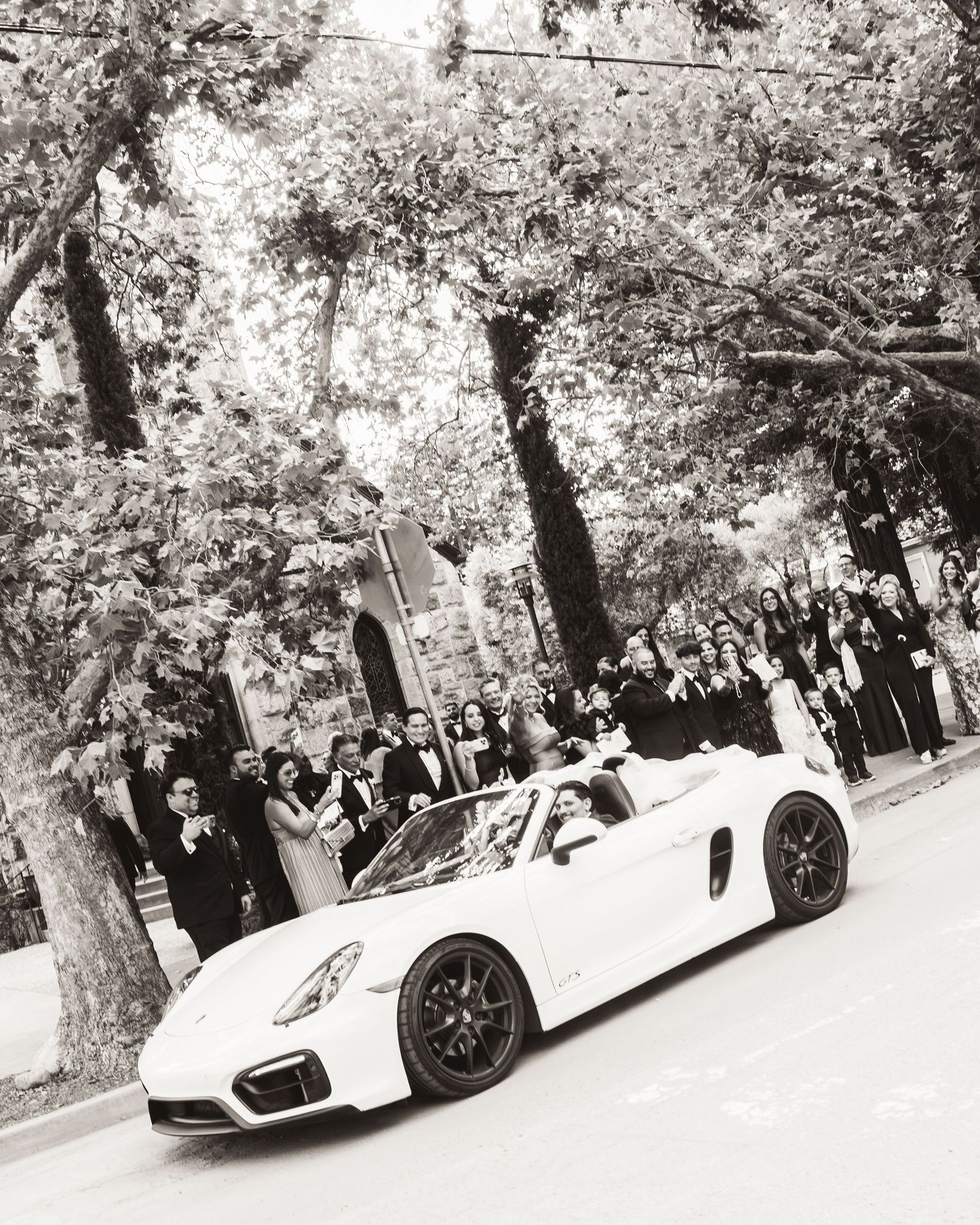 Bride and groom departing wedding in white Porsche convertible as guests celebrate.