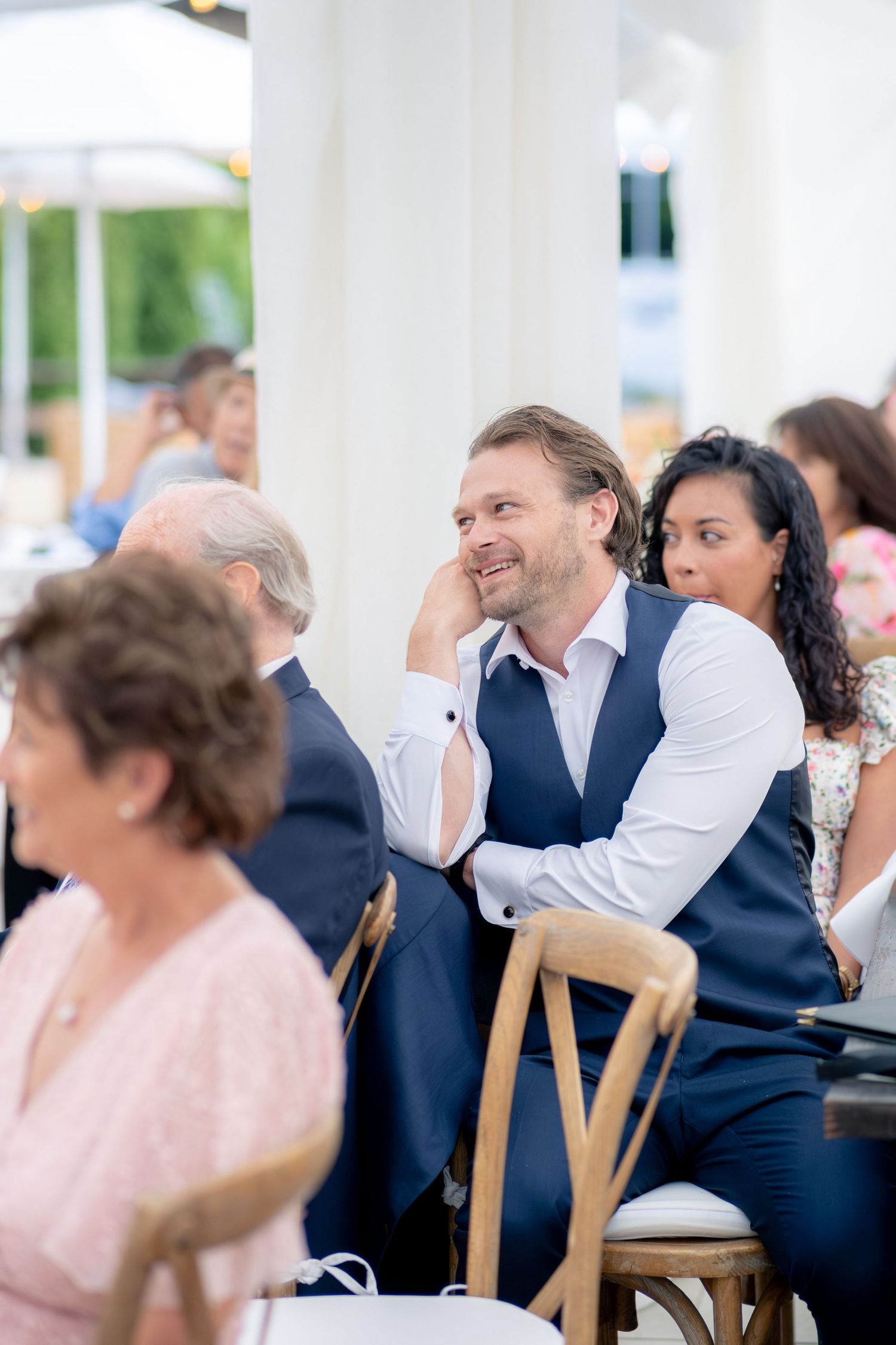 Smiling male wedding guest resting chin on hand while watching ceremony under white tent.