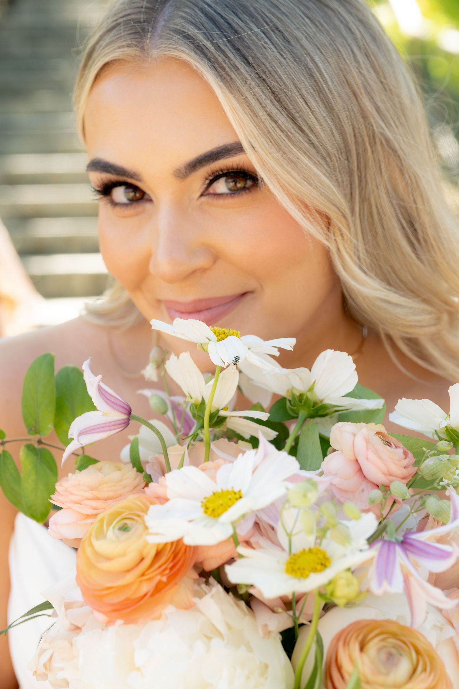 Close-up portrait of blonde bride smiling with a pastel bouquet of ranunculus, daisies, and wildflowers.