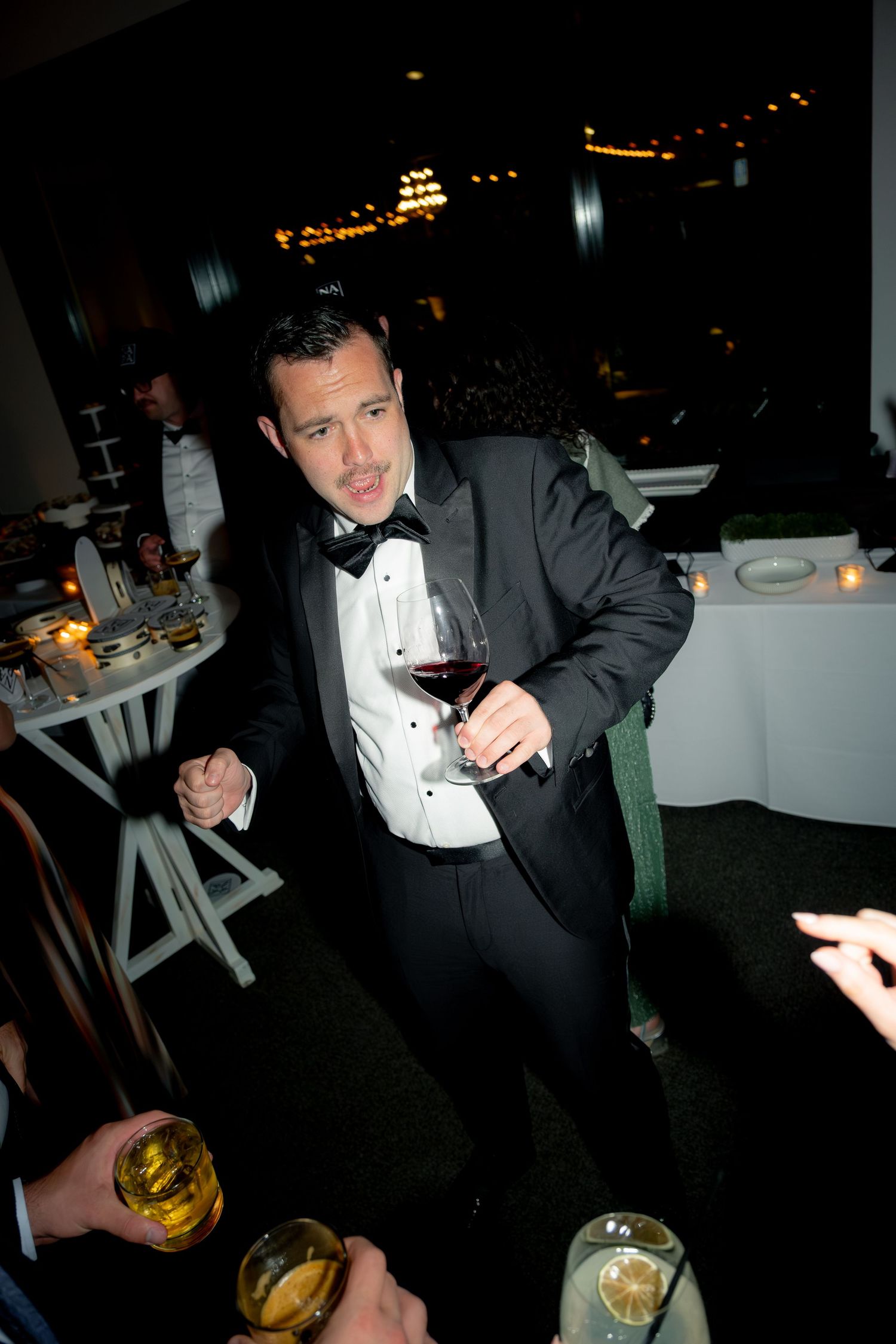 Dapper groomsman in tuxedo holds red wine glass while socializing at elegant evening wedding reception.