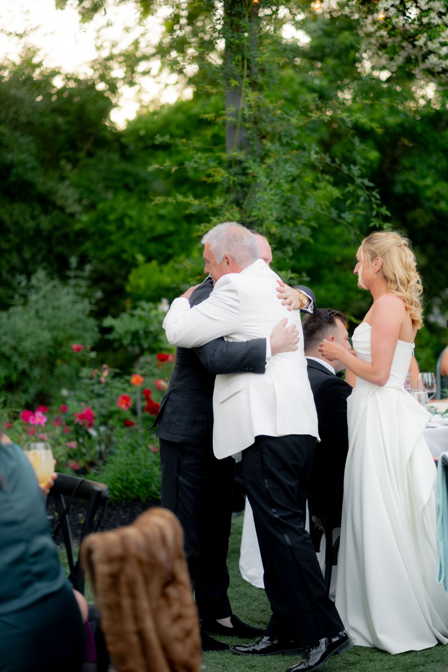 Groom in white jacket embraces guest while bride in white gown watches at outdoor garden wedding reception.