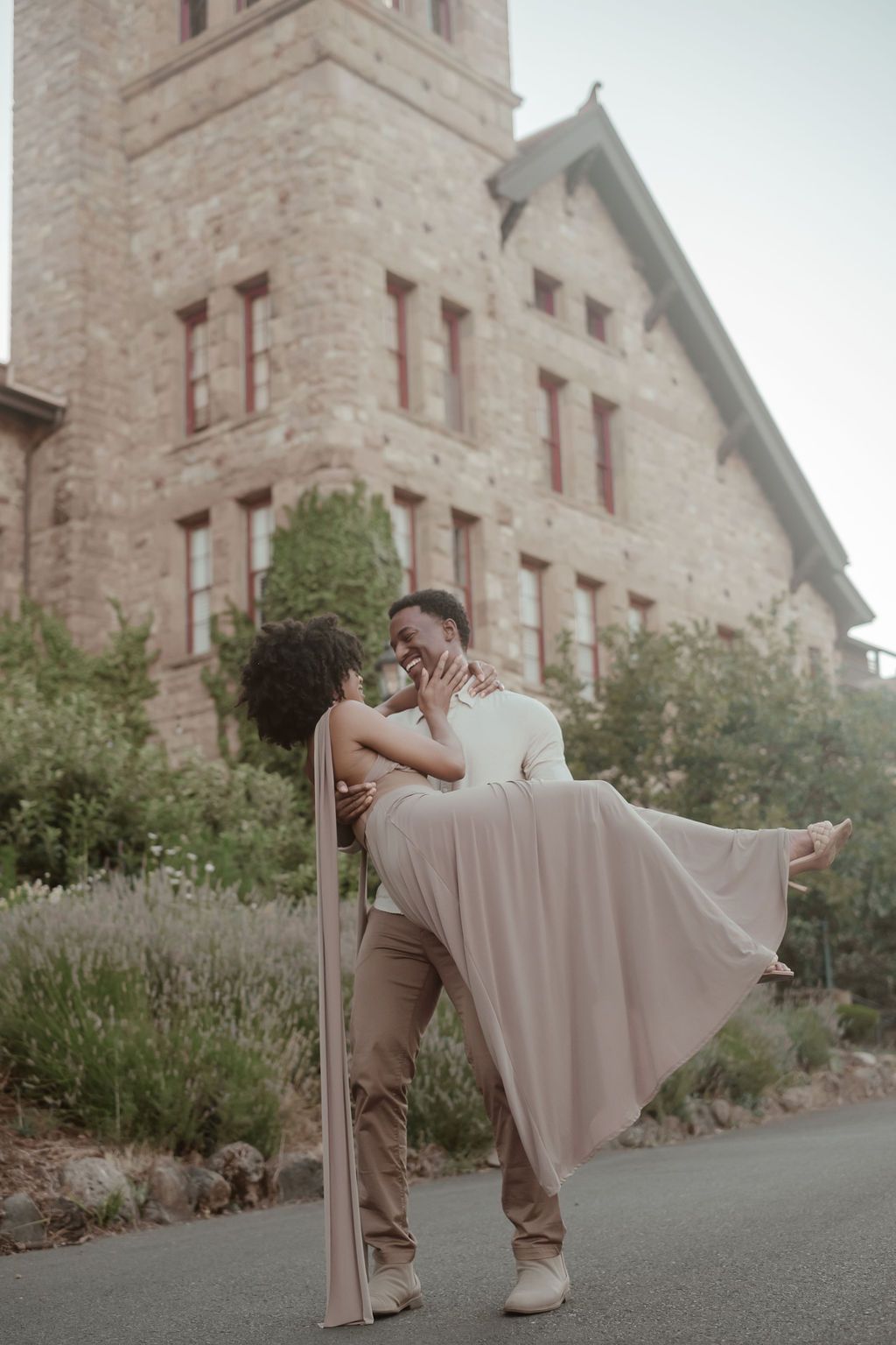 A joyful man lifts his partner in a flowing mauve gown outside a historic stone building.