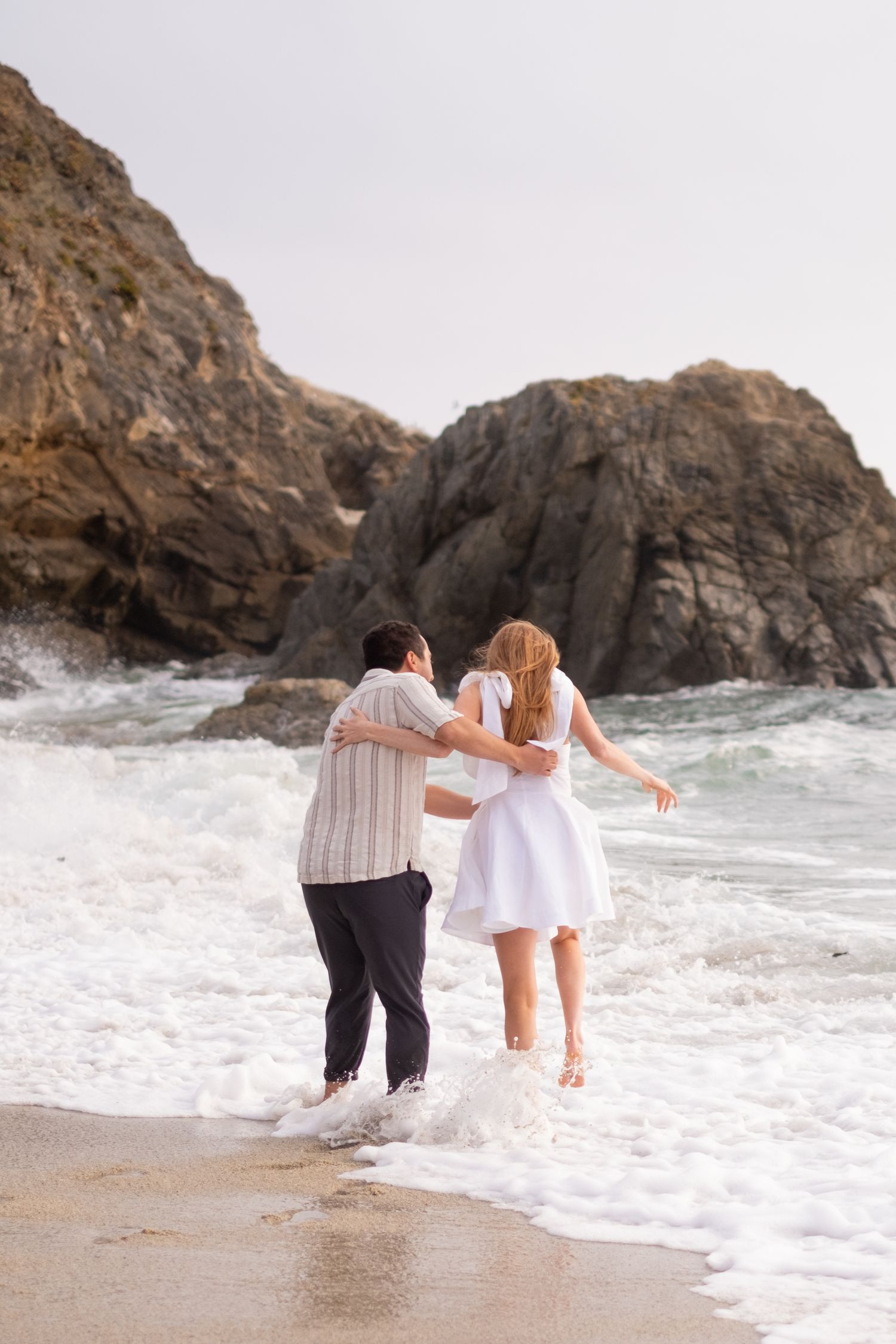 Couple embracing in ocean waves on a rocky beach, woman in white dress and man in striped shirt.