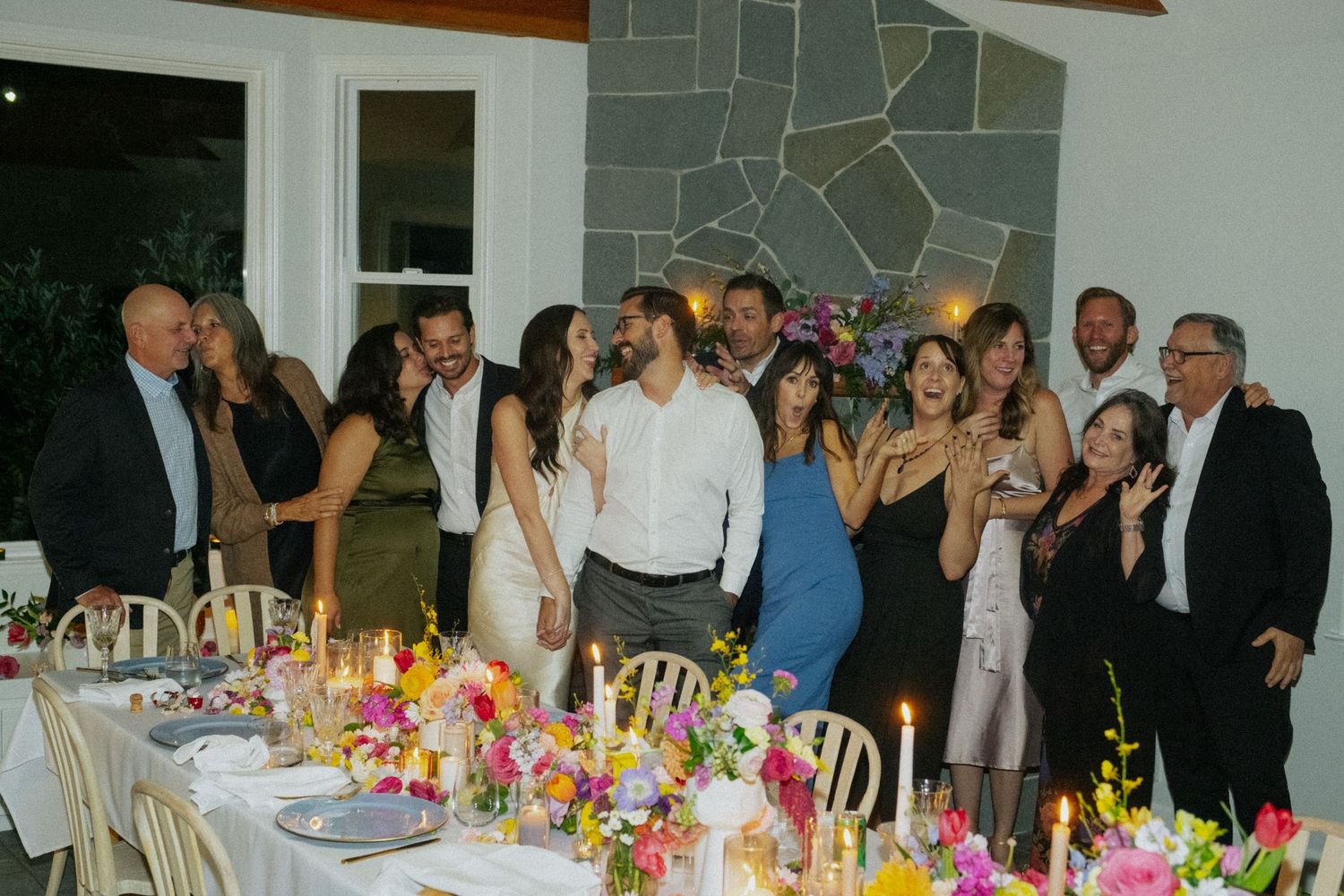 Wedding reception group photo with guests gathered around a candlelit table adorned with colorful floral arrangements.