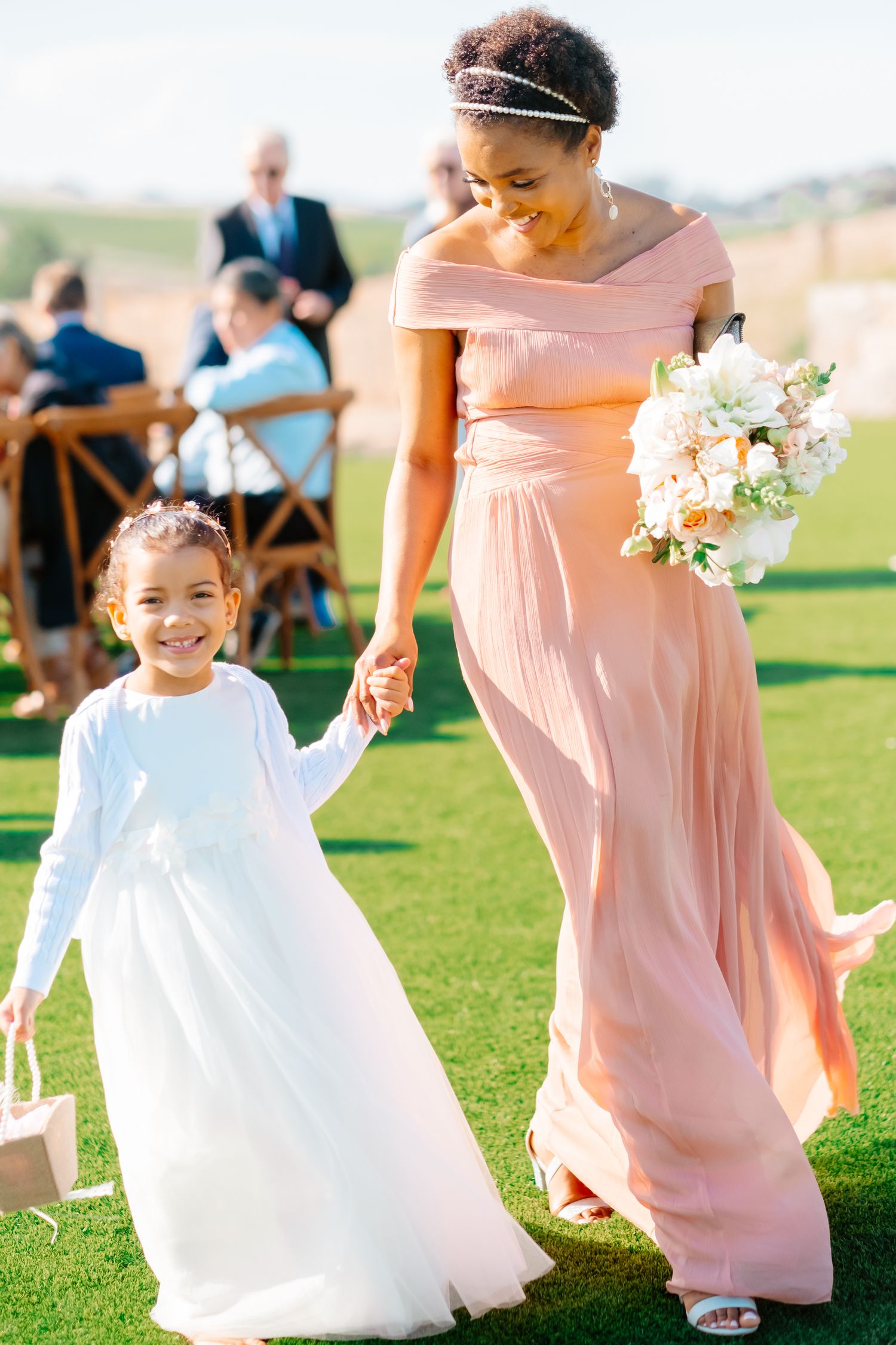 Bridesmaid in blush gown walks smiling flower girl in white dress down green grass aisle.