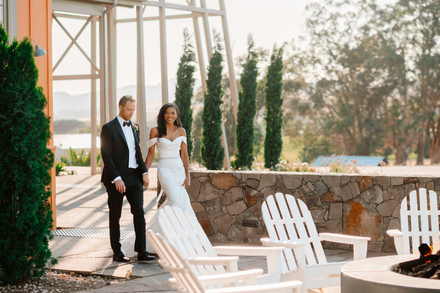 Bride in fitted white gown and groom in black tuxedo walking together at golden hour outdoor venue.