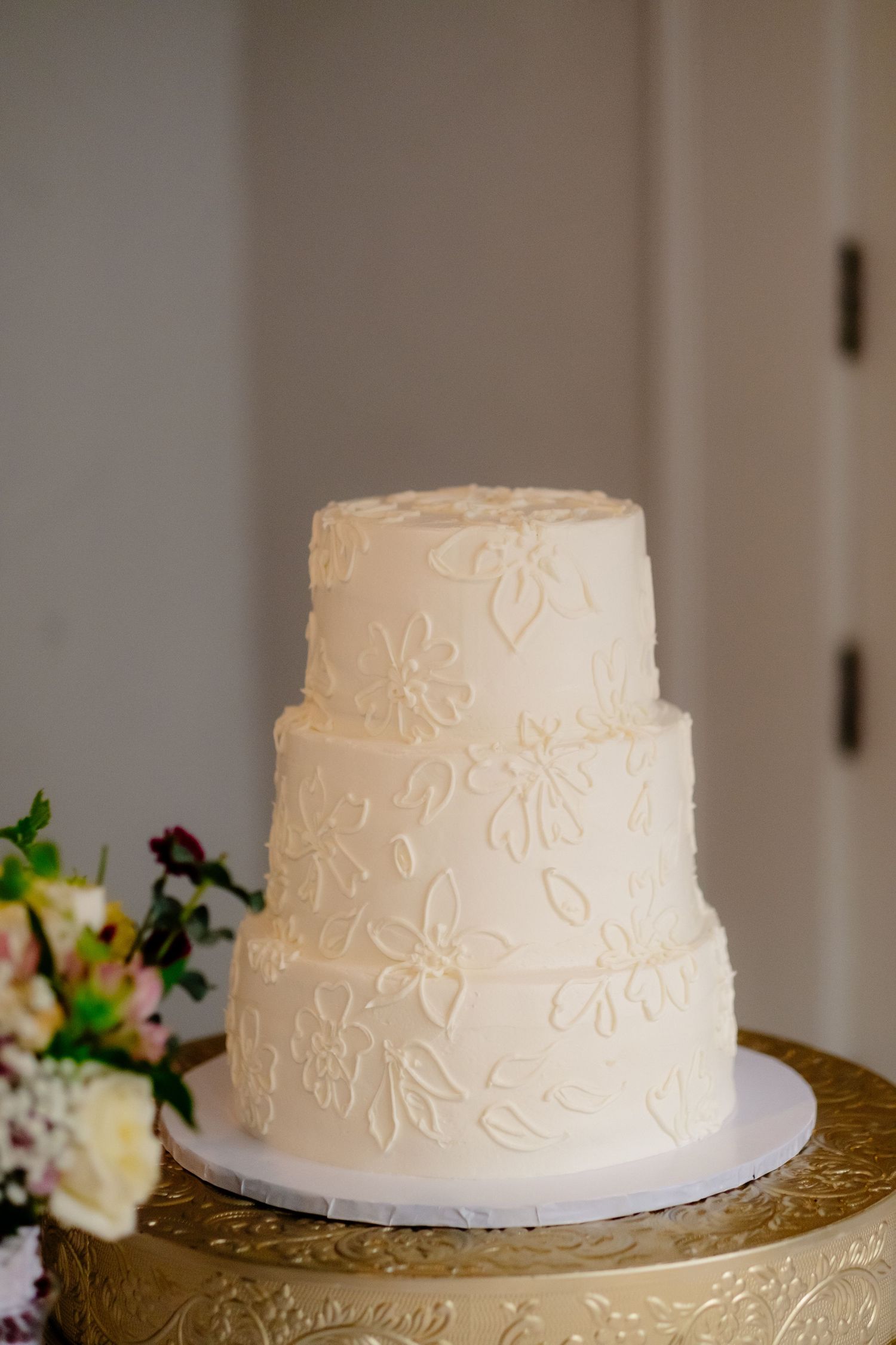 Three-tier white wedding cake with floral piping detail on gold cake stand.