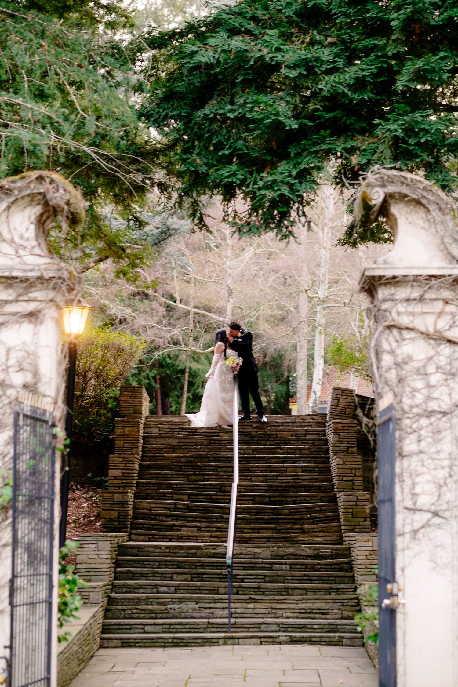 Bride and groom kissing at the top of stone stairs framed by ornate gates and trees.