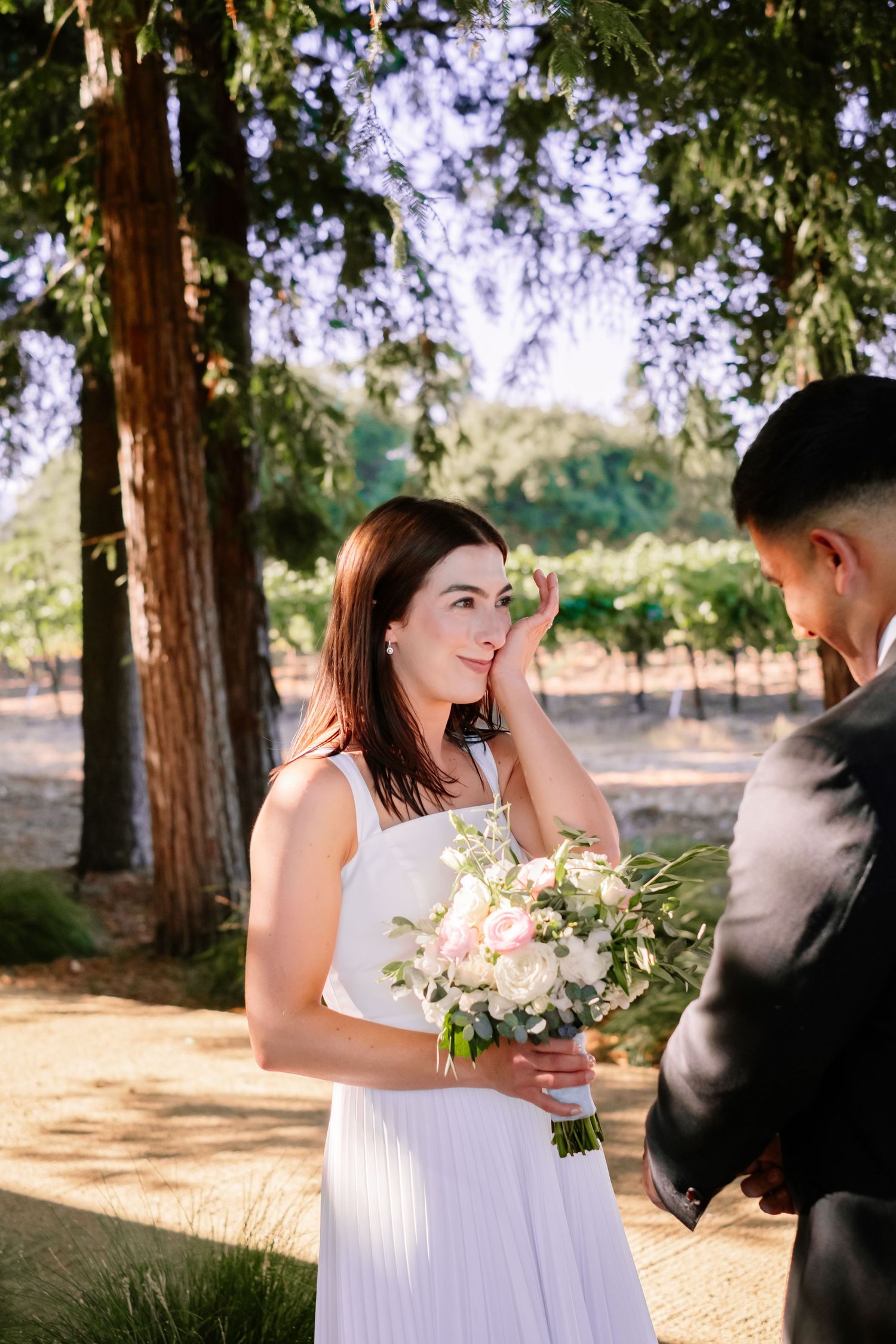 Emotional bride in white pleated dress holding pink bouquet wiping tear during outdoor ceremony.