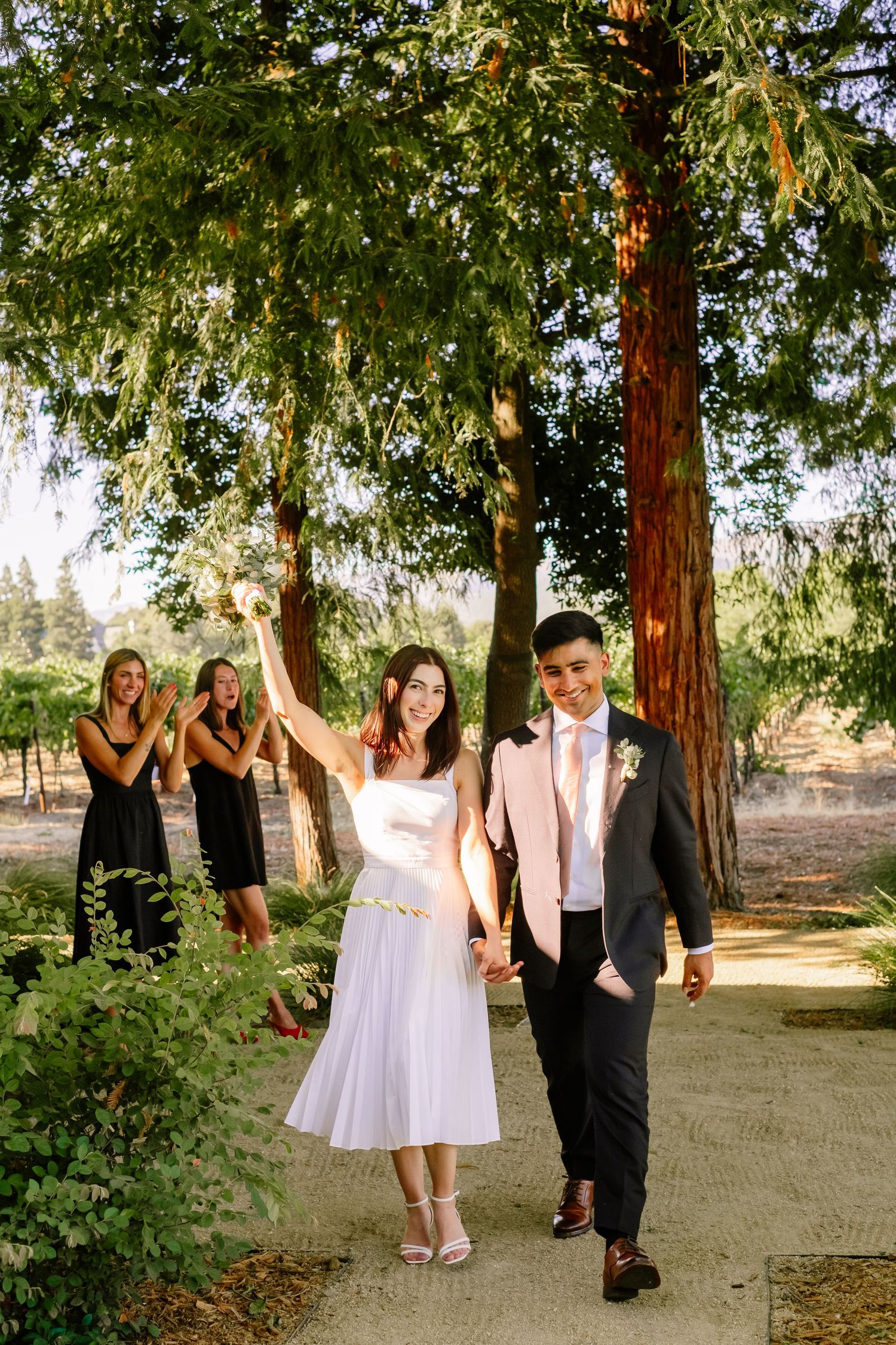 Joyful bride raises bouquet while walking with groom through sunlit redwood tree path.