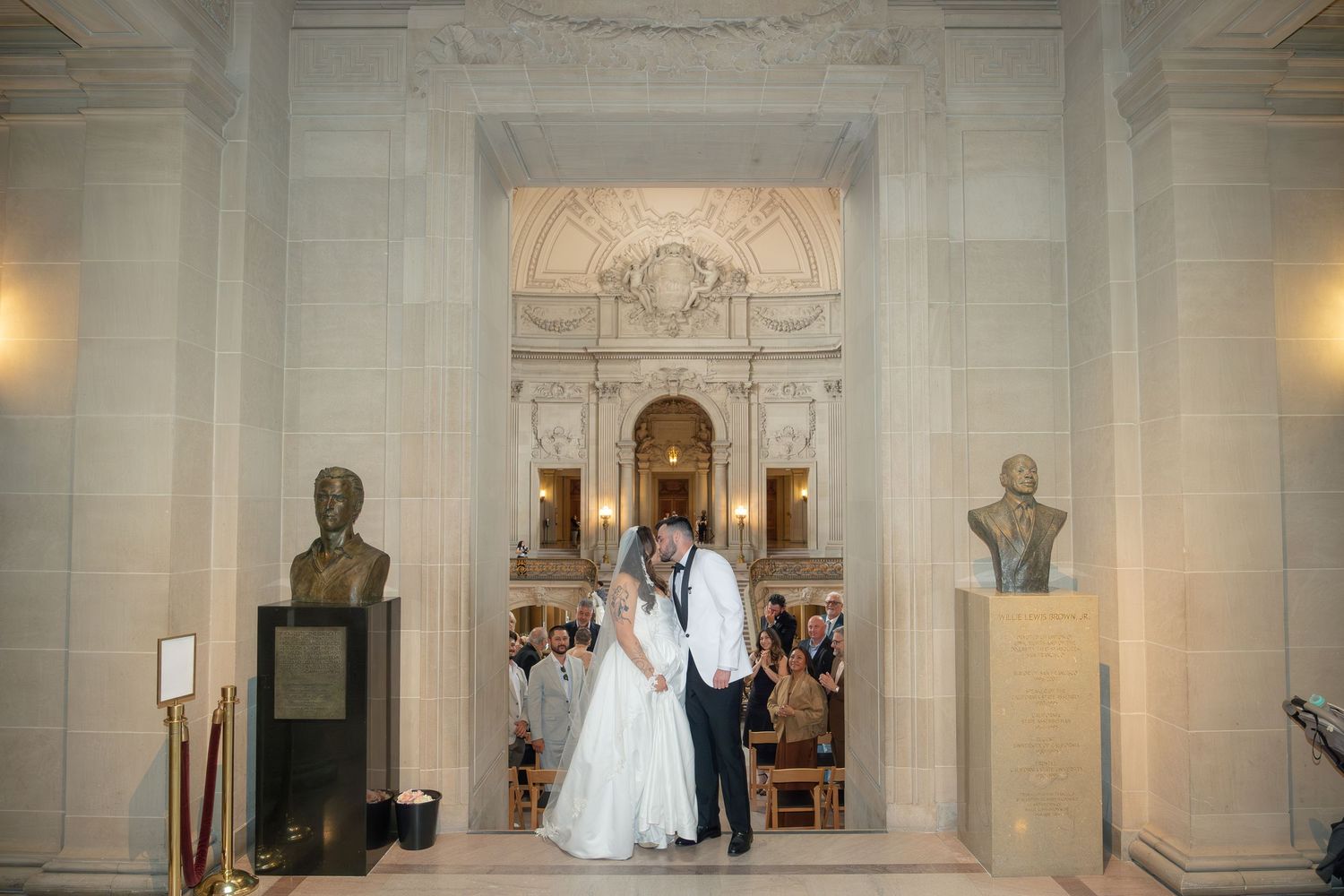 Bride and groom share a kiss inside an elegant marble hall with busts and arched alcoves at their wedding ceremony.