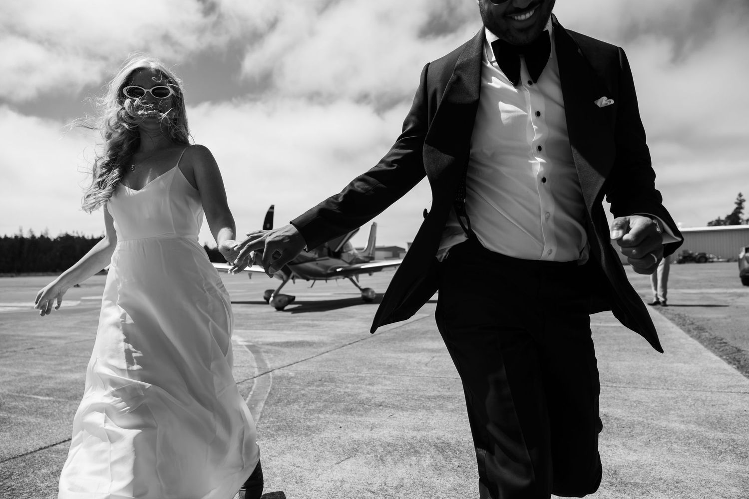 Black and white photo of a bride and groom holding hands walking on an airstrip with a small plane in the background.