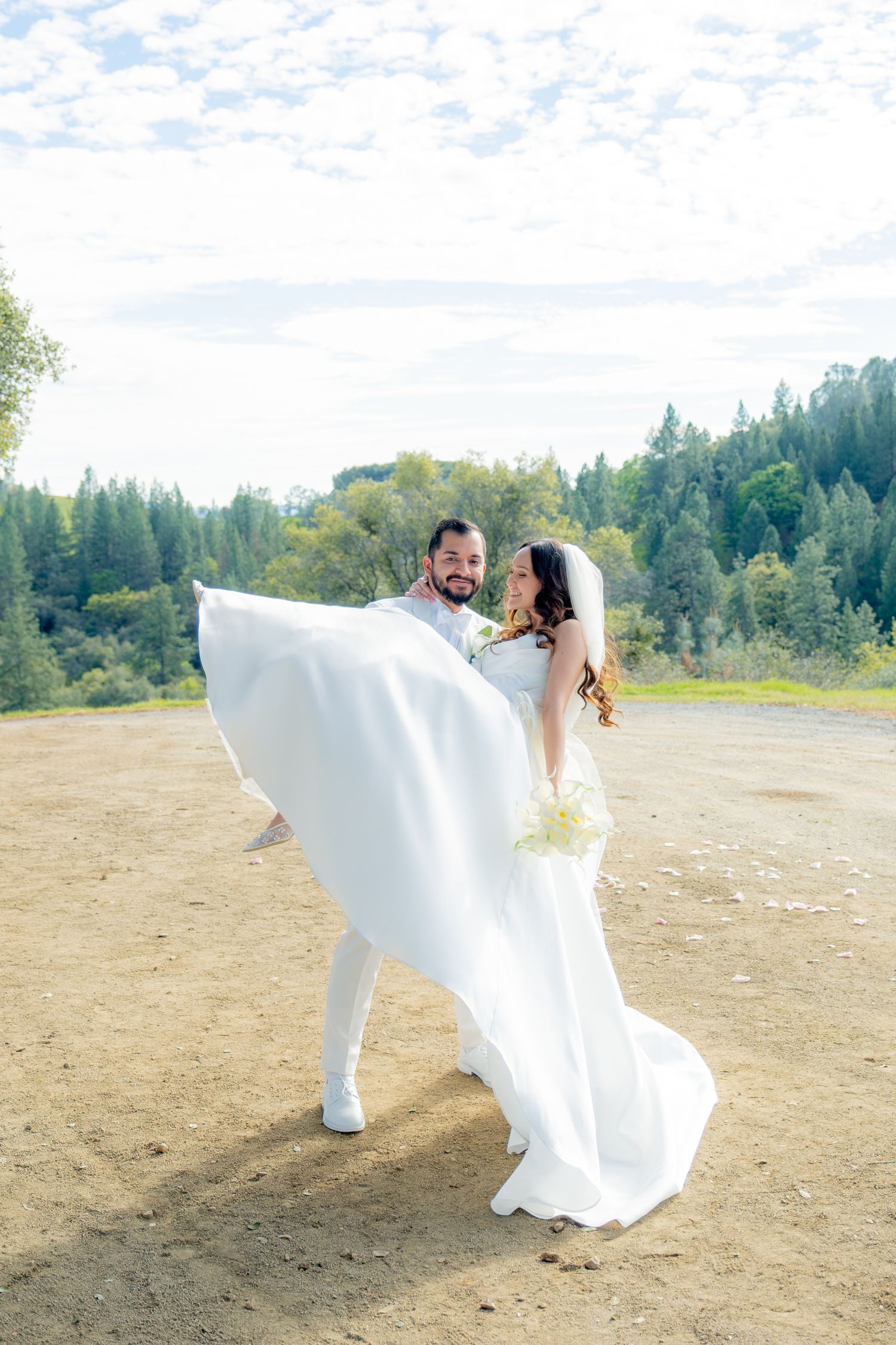 Joyful newlyweds embracing outdoors with bride's veil flowing in the wind against a forest backdrop.