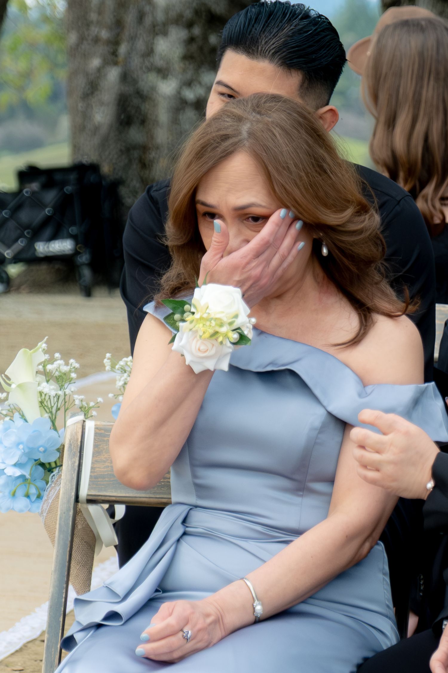 Emotional mother of the bride in blue dress wiping tears during outdoor wedding ceremony.