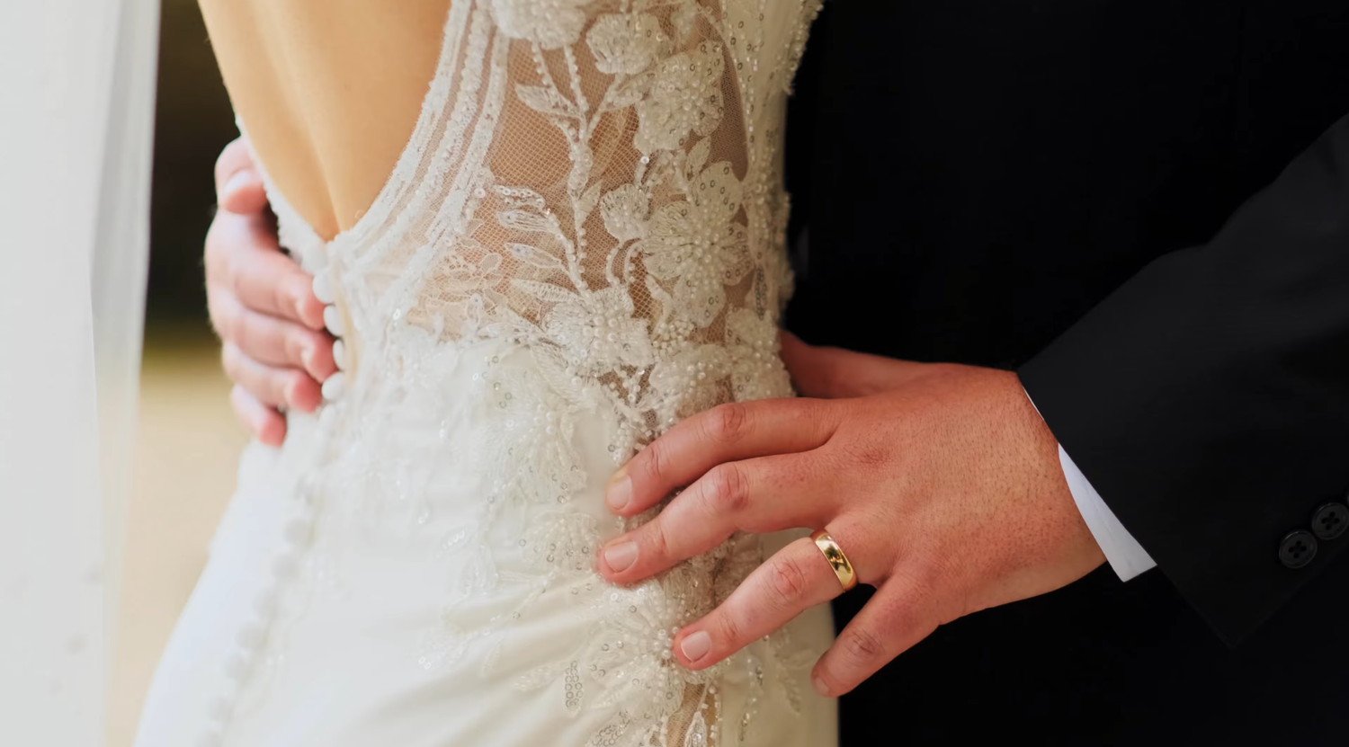 Close-up of groom placing hand on bride's intricate lace wedding dress back detail.