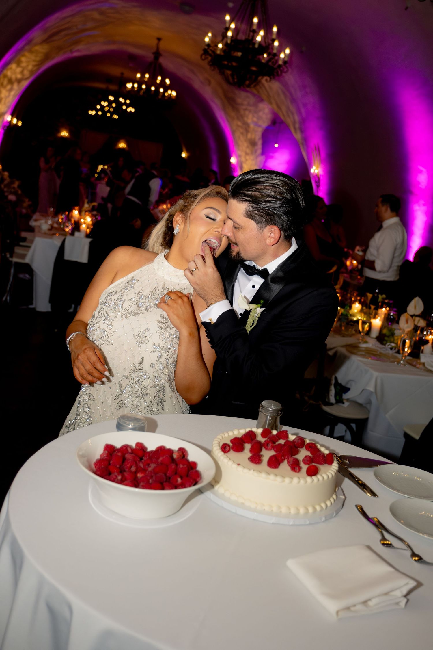 Bride and groom sharing cake at elegant wedding reception with purple uplighting.