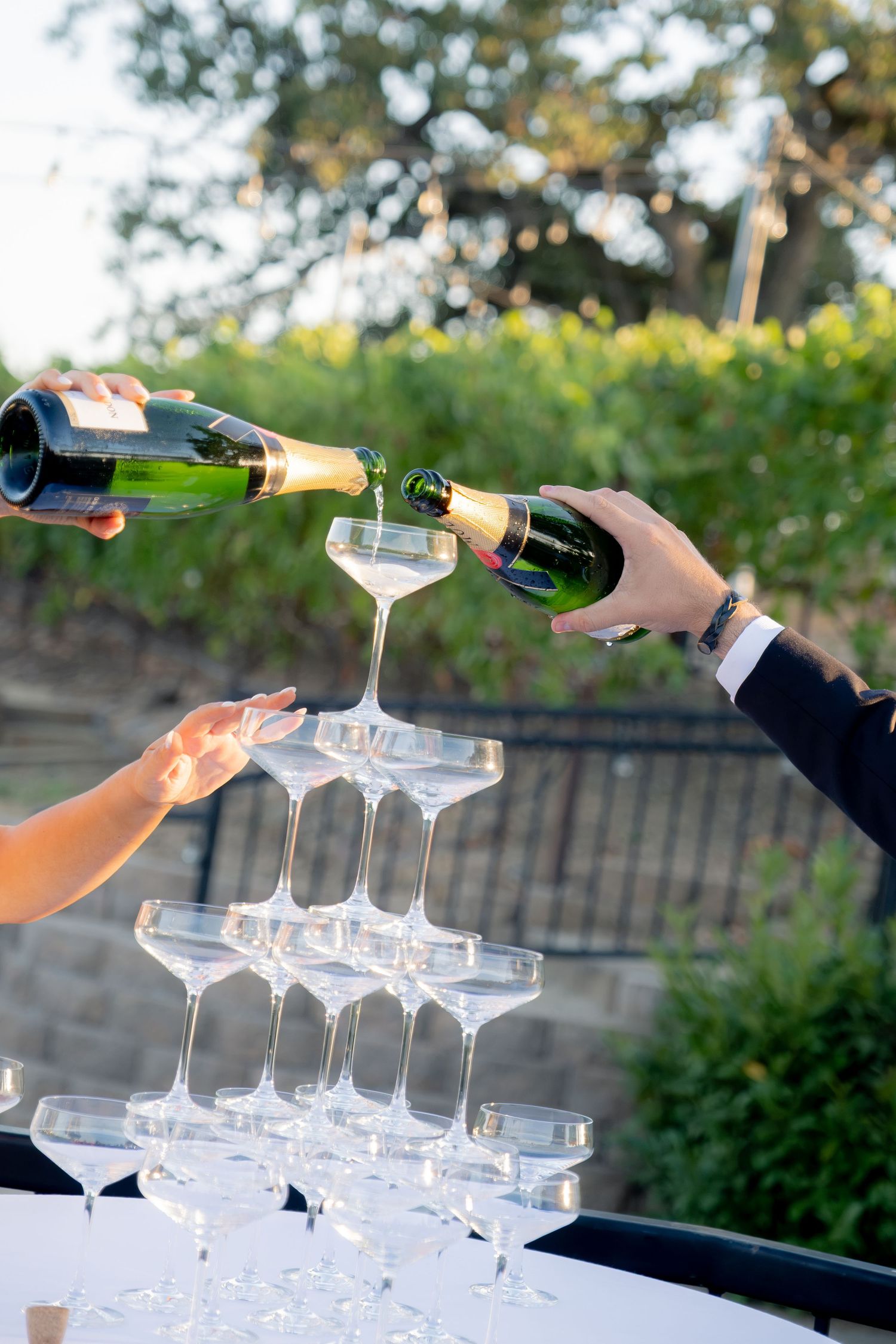 Two people pouring champagne into a elegant tower of coupe glasses at an outdoor wedding reception.