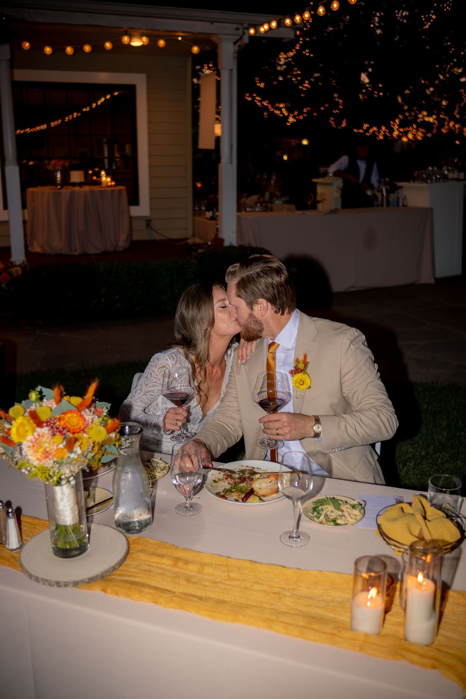 Newlyweds sharing a kiss at their outdoor reception dinner table adorned with vibrant floral centerpieces at night.