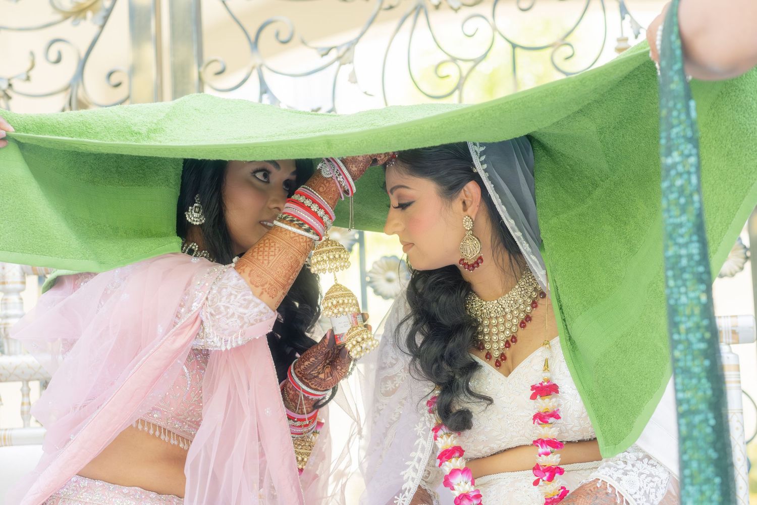 Two brides sharing an intimate moment under a green dupatta at an Indian wedding.