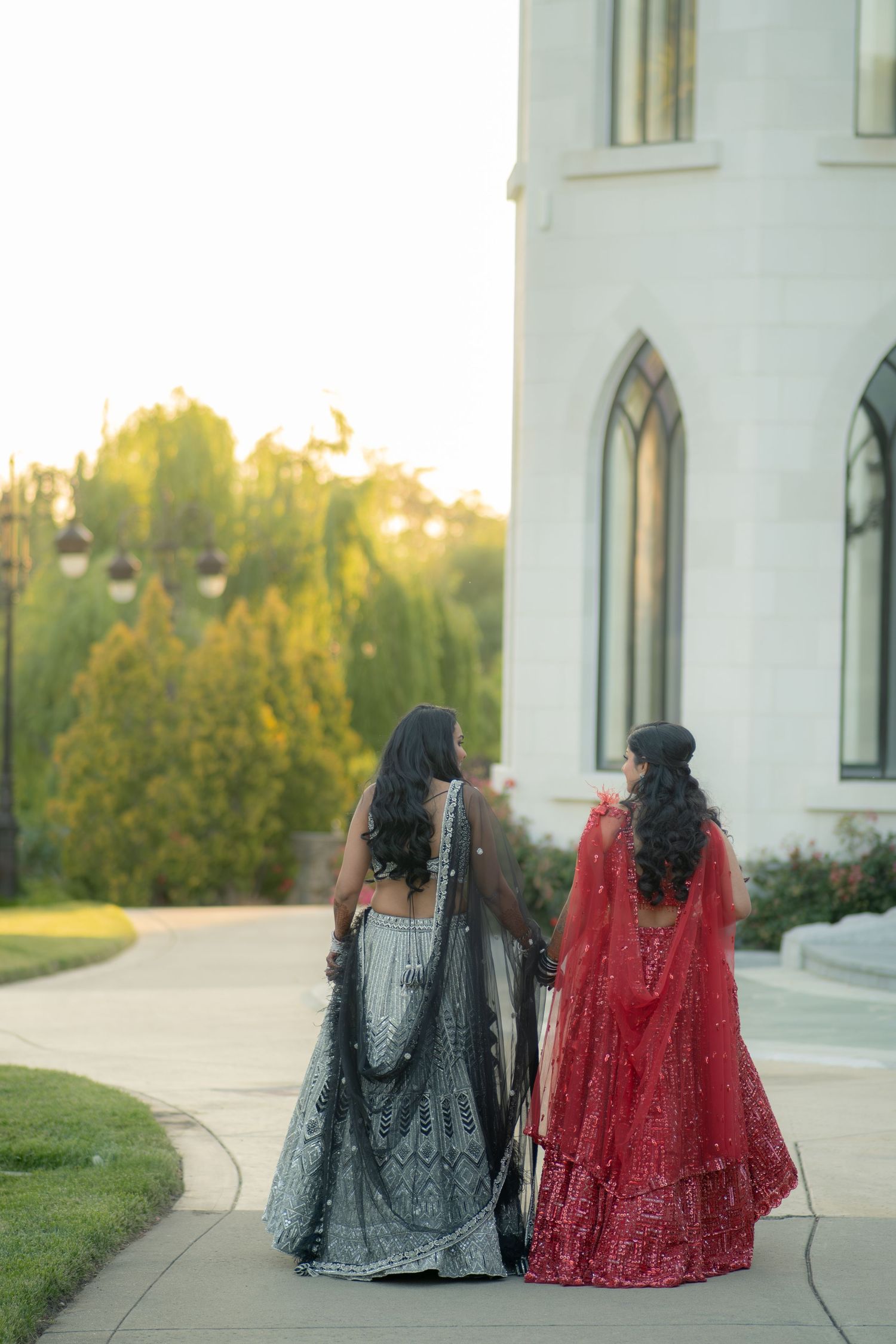Two women in red and grey Indian lehengas walk hand in hand toward a white building at golden hour.