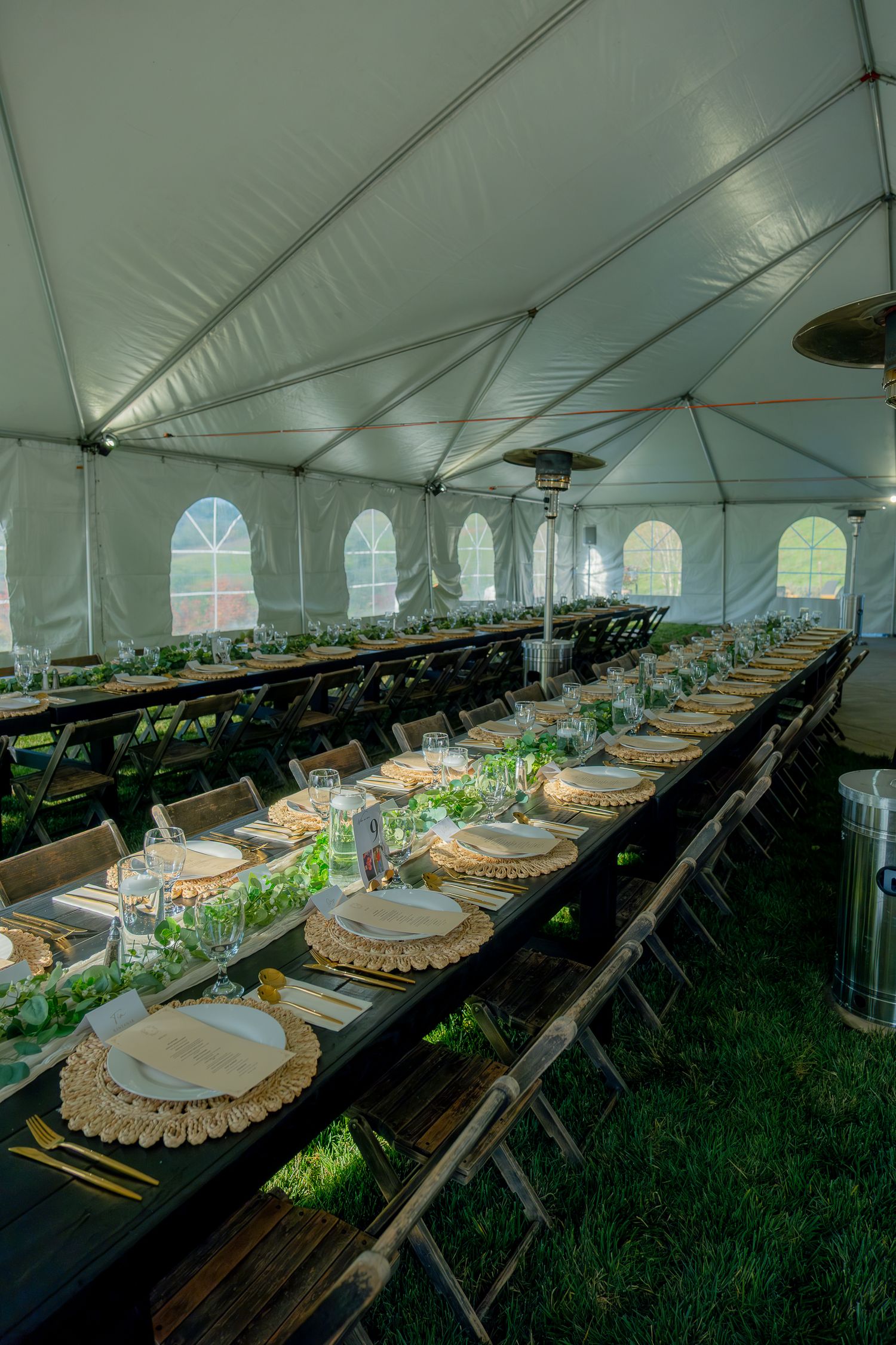 Elegant wedding reception tent with long dark tables, greenery centerpieces, and woven chargers.