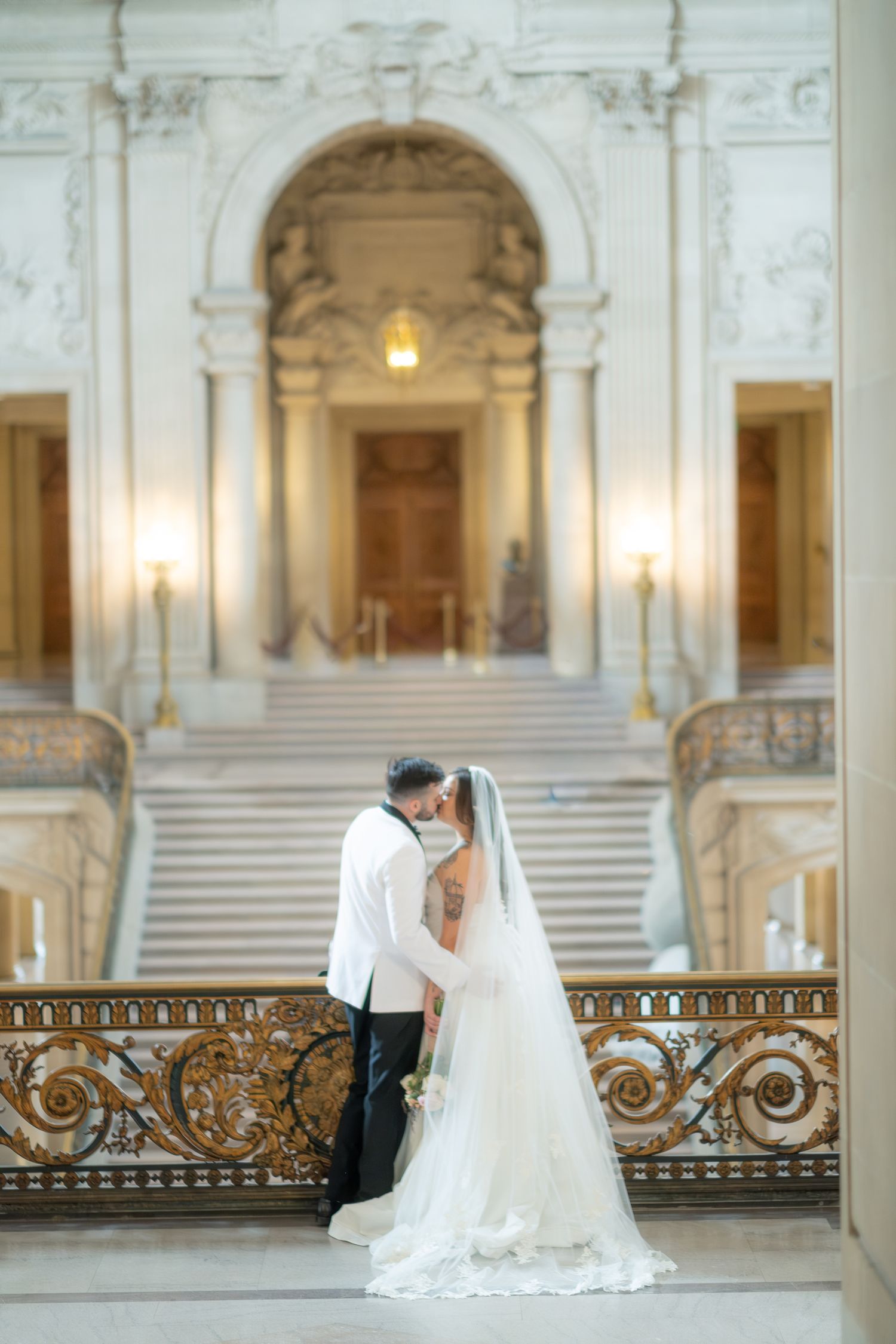 Newlyweds share a kiss on the grand balcony of San Francisco City Hall, bride in flowing white gown with veil.