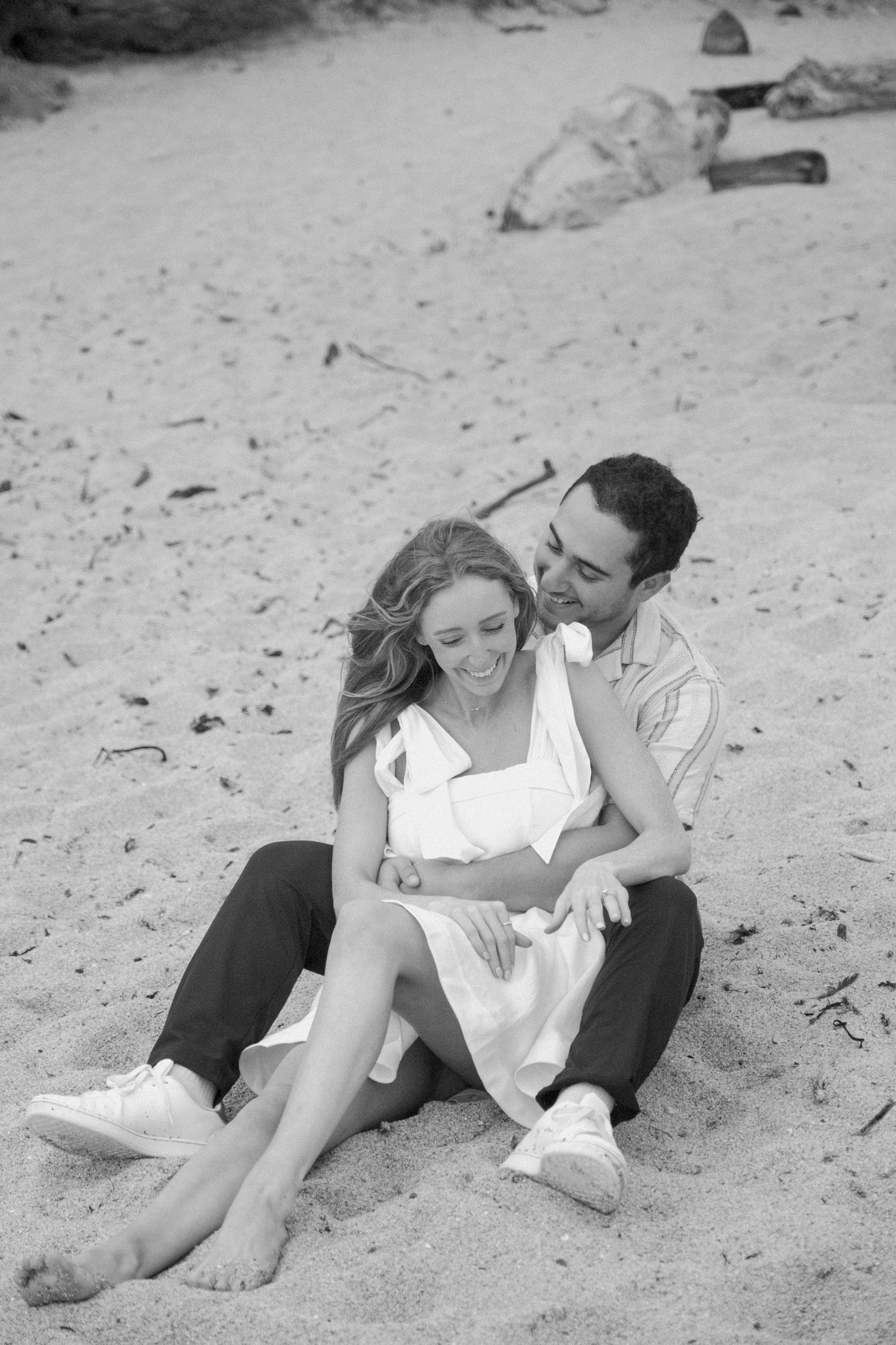 Black and white photo of couple sitting together on sandy beach, woman in white dress laughing joyfully.