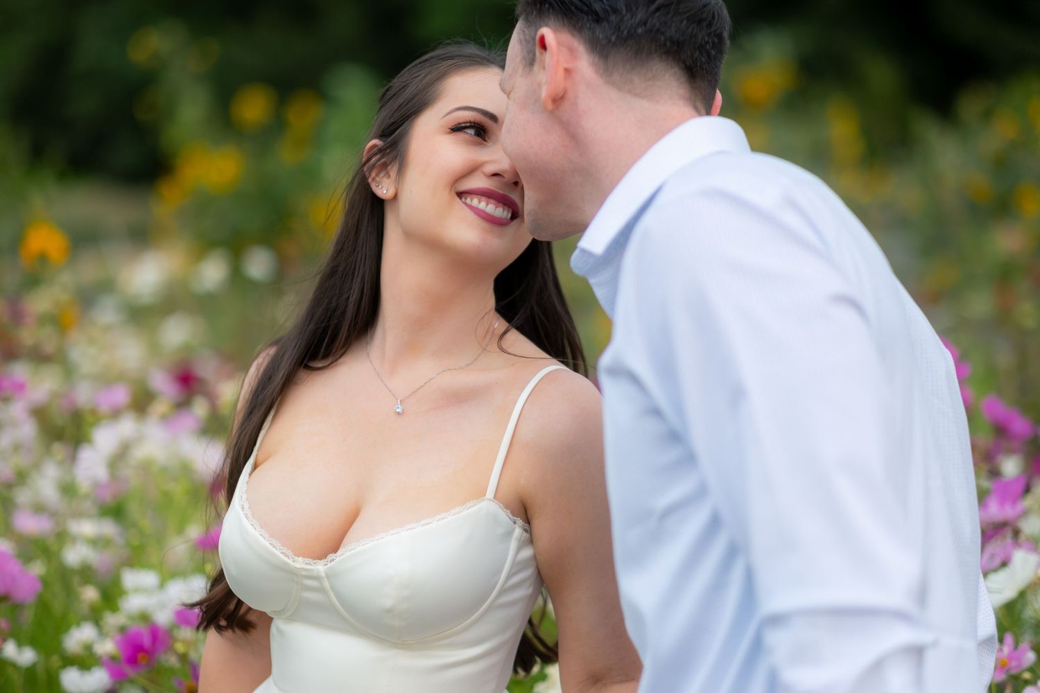 Smiling bride in white dress with groom whispering in flower field.