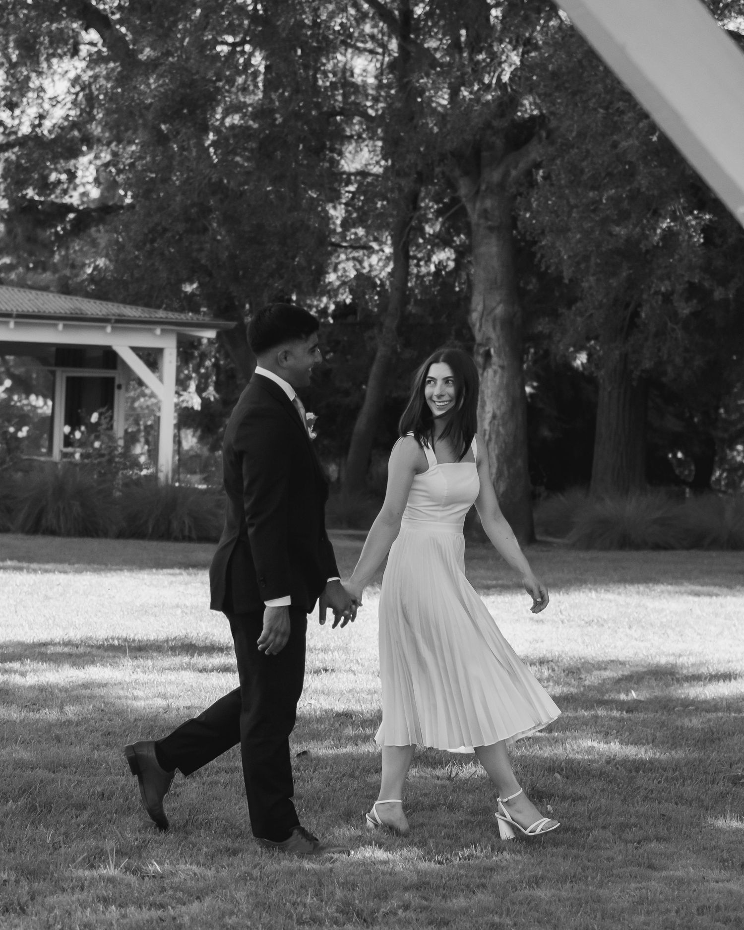 Black and white photo of a couple holding hands, walking on grass, woman in white dress, man in dark suit.