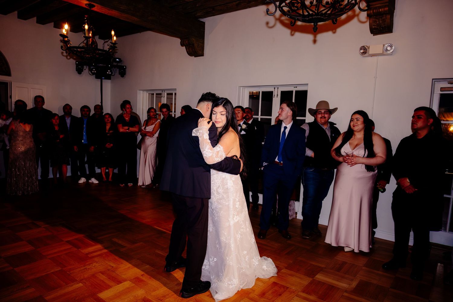 Bride and groom share romantic first dance on wooden dance floor surrounded by wedding guests.