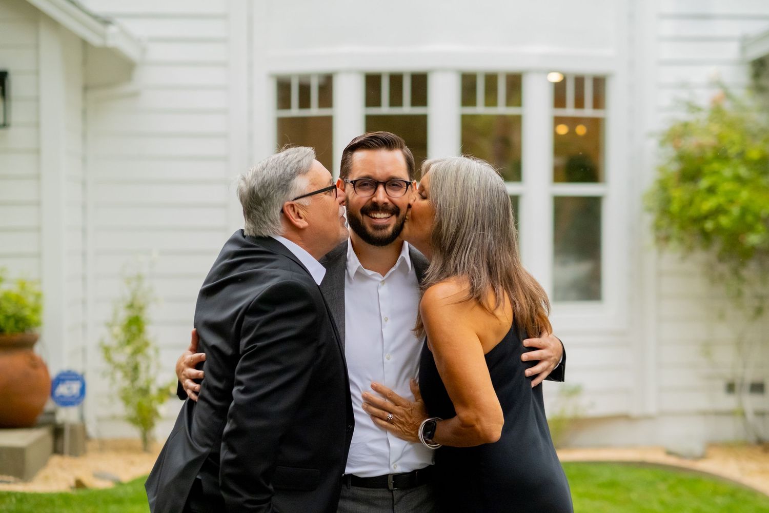 A joyful man receives kisses on the cheek from two people outside a white building.