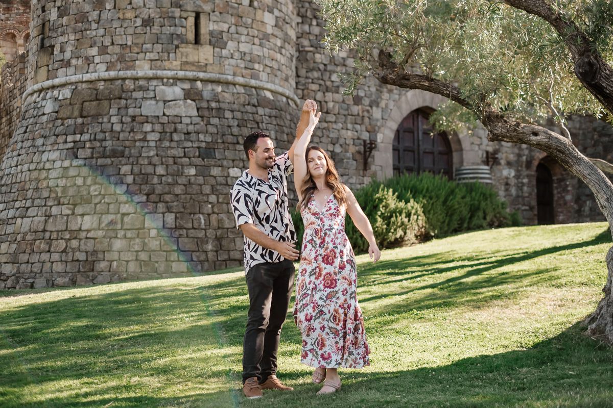 Joyful engaged couple dancing on grass beside a stone castle tower with olive tree.